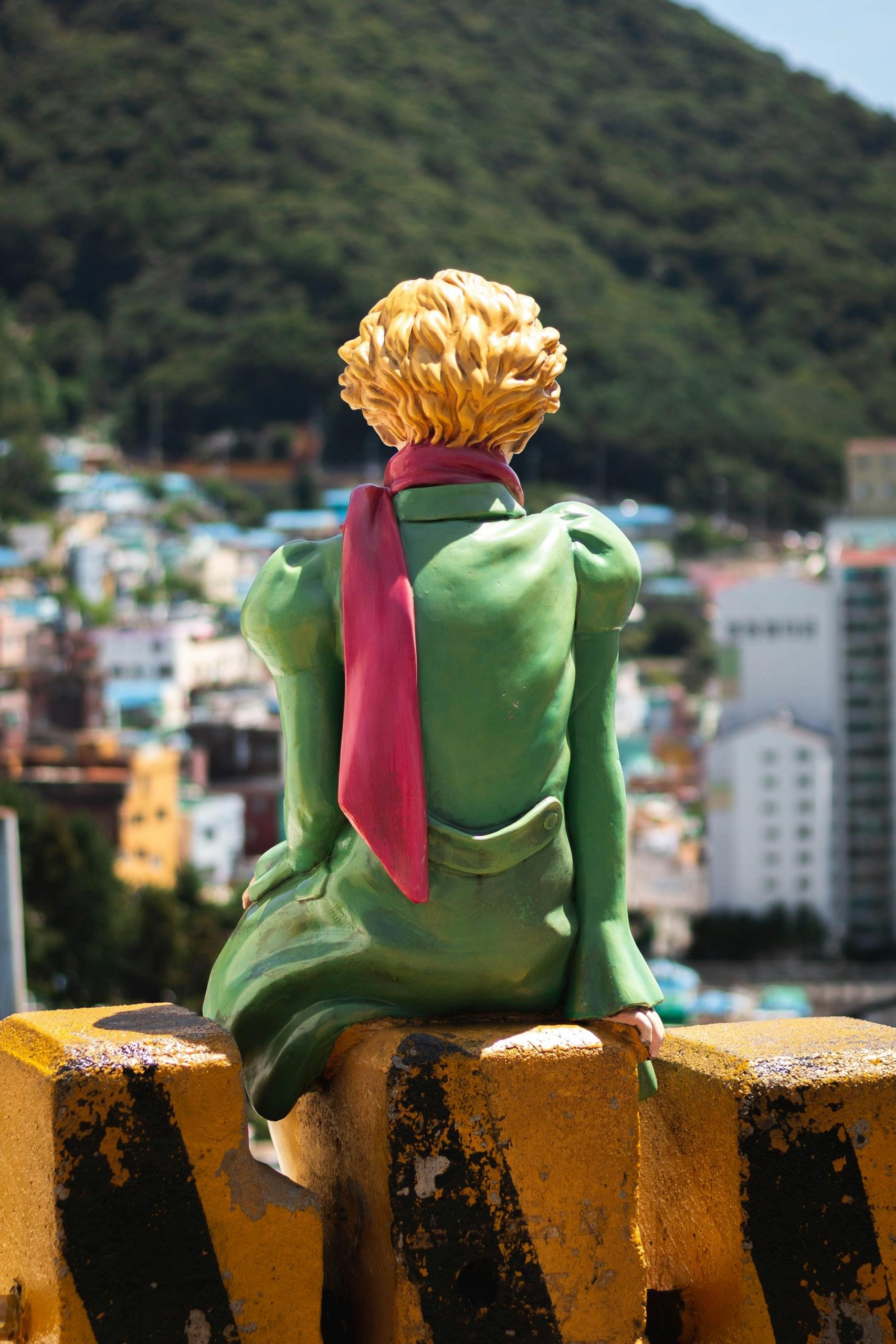 Una estatua con abrigo verde y bufanda roja sentada en una barrera, con vistas a un paisaje urbano montañoso.