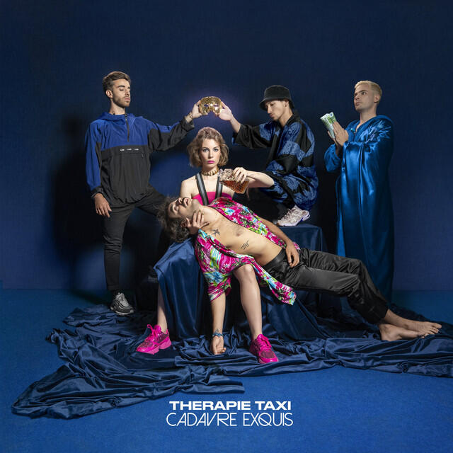 La banda «Therapie Taxi» posa en un escenario oscuro y dramático. Una mujer está sentada, sosteniendo la cabeza de un hombre recostado. Otros sostienen accesorios, creando una escena surrealista y artística.