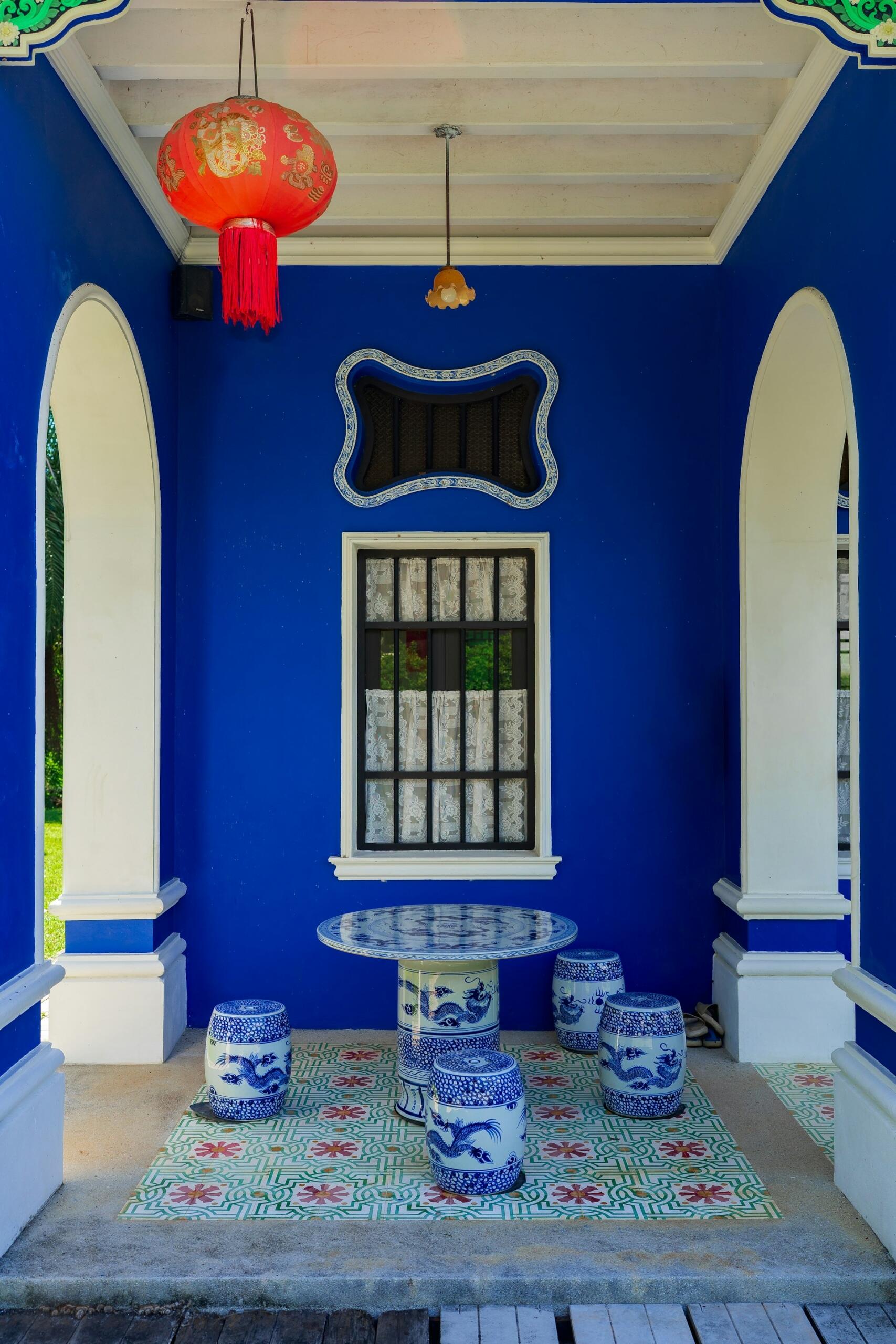 Patio cubierto con paredes de un azul intenso, suelo de baldosas ornamentadas y mesa circular con taburetes a juego. Una linterna roja cuelga del techo, creando un ambiente tranquilo y decorativo.