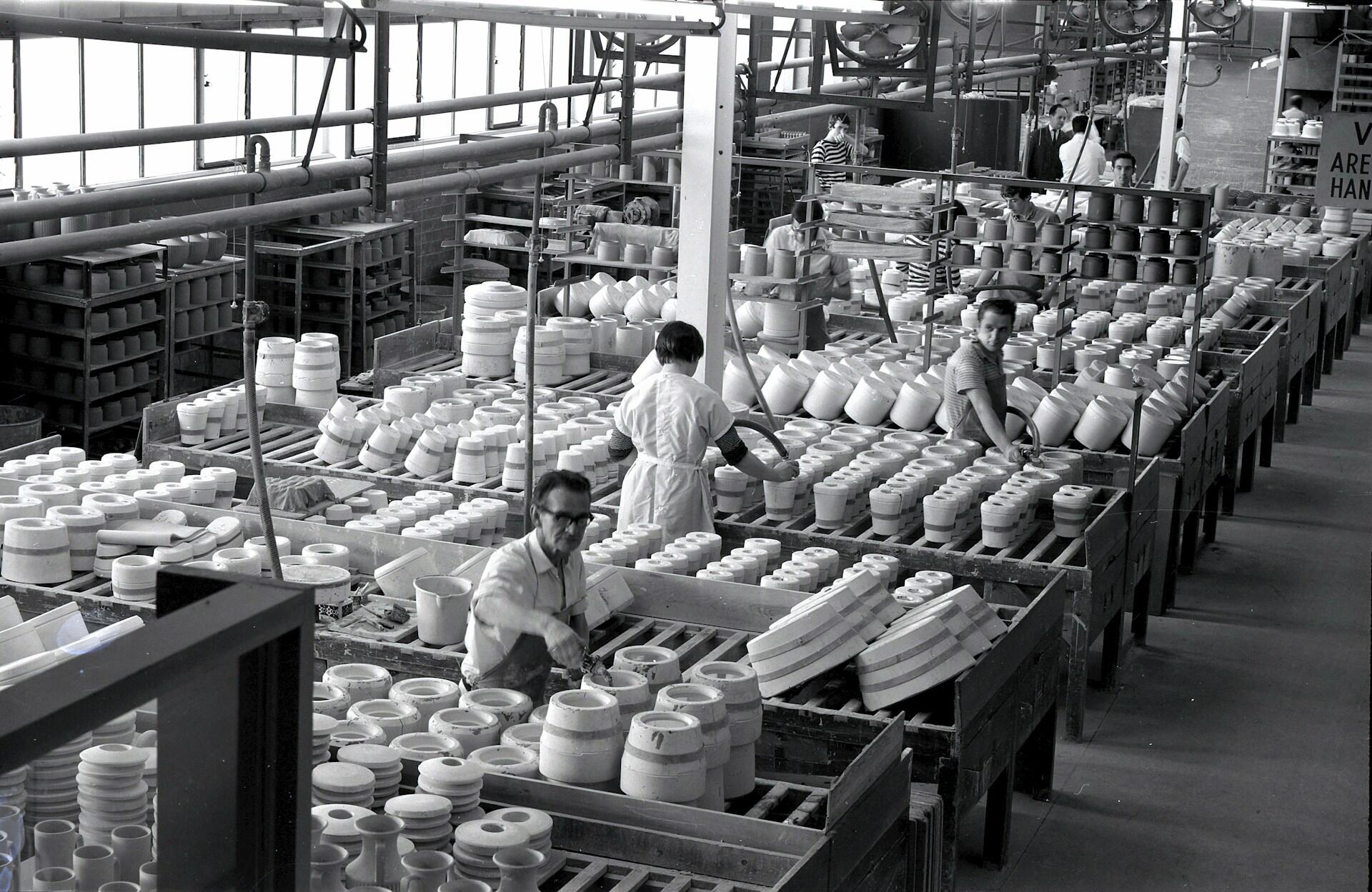 Imagen en blanco y negro de una bulliciosa fábrica de cerámica. Trabajadores uniformados manipulan filas de piezas de cerámica sobre largas mesas. Ambiente industrial, organizado y ajetreado.