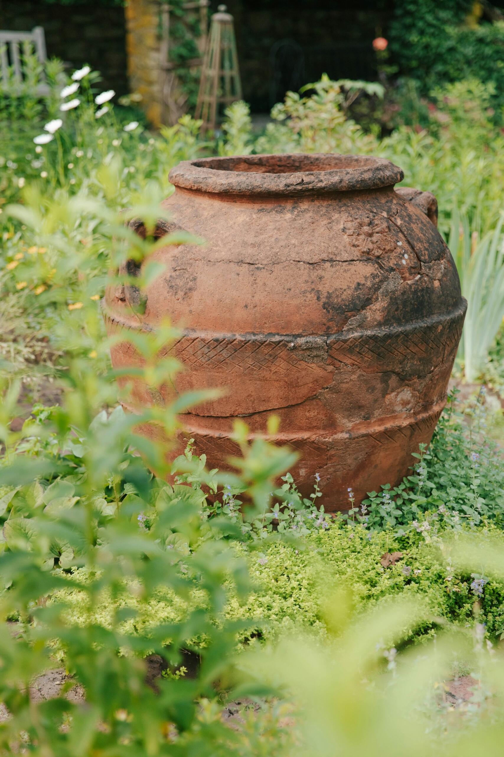 Una gran urna de terracota desgastada por el paso del tiempo se encuentra en medio de la exuberante vegetación de un jardín. La suave luz del sol y las flores en flor evocan una atmósfera serena y atemporal.