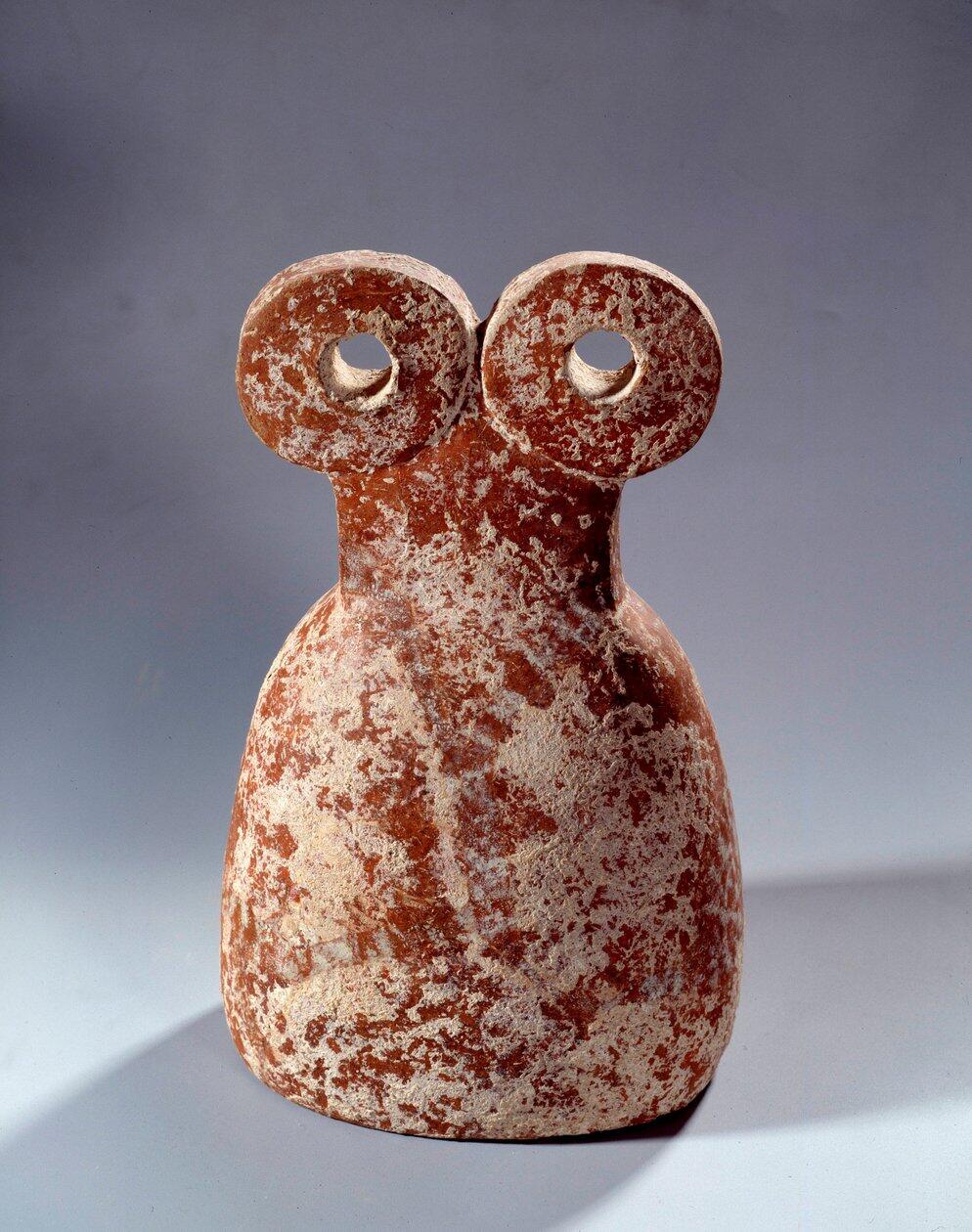 Una pequeña escultura de mármol con forma de búho y una textura suave y desgastada. La figura tiene dos grandes ojos circulares y un cuerpo redondeado, lo que le da un aspecto caprichoso.