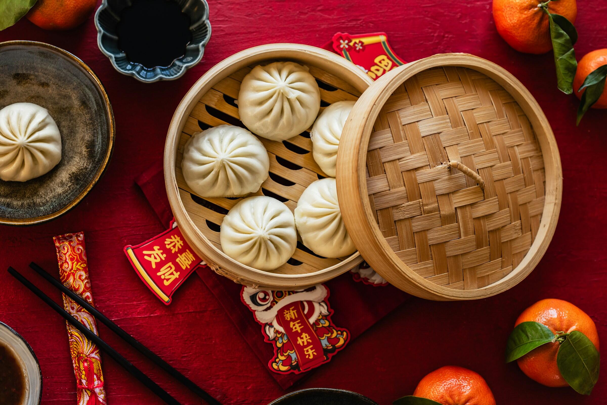 bollos chinos al vapor dispuestos en una cesta de bambú tradicional. El fondo rojo y los elementos decorativos evocan una celebración cultural asiática.
