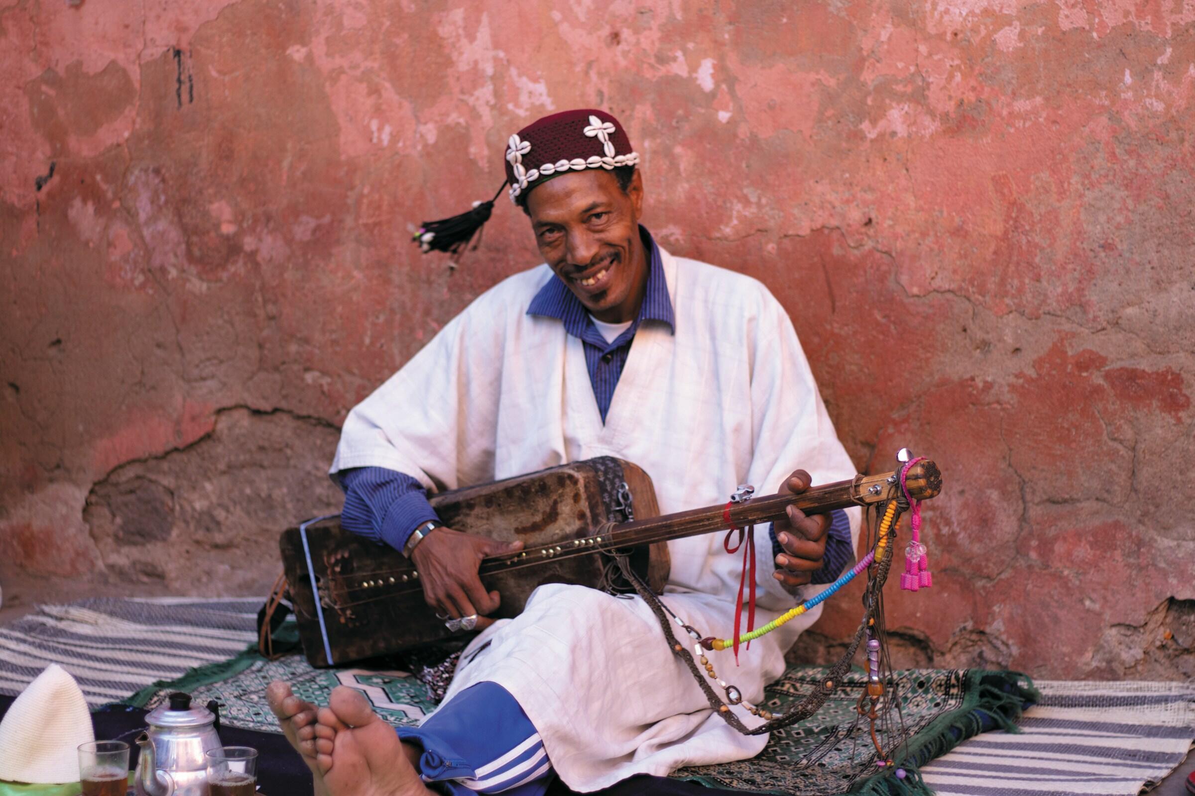 Hombre vestido con traje tradicional tocando un instrumento de cuerda, sentado sobre alfombras estampadas frente a una pared con textura y aspecto desgastado.