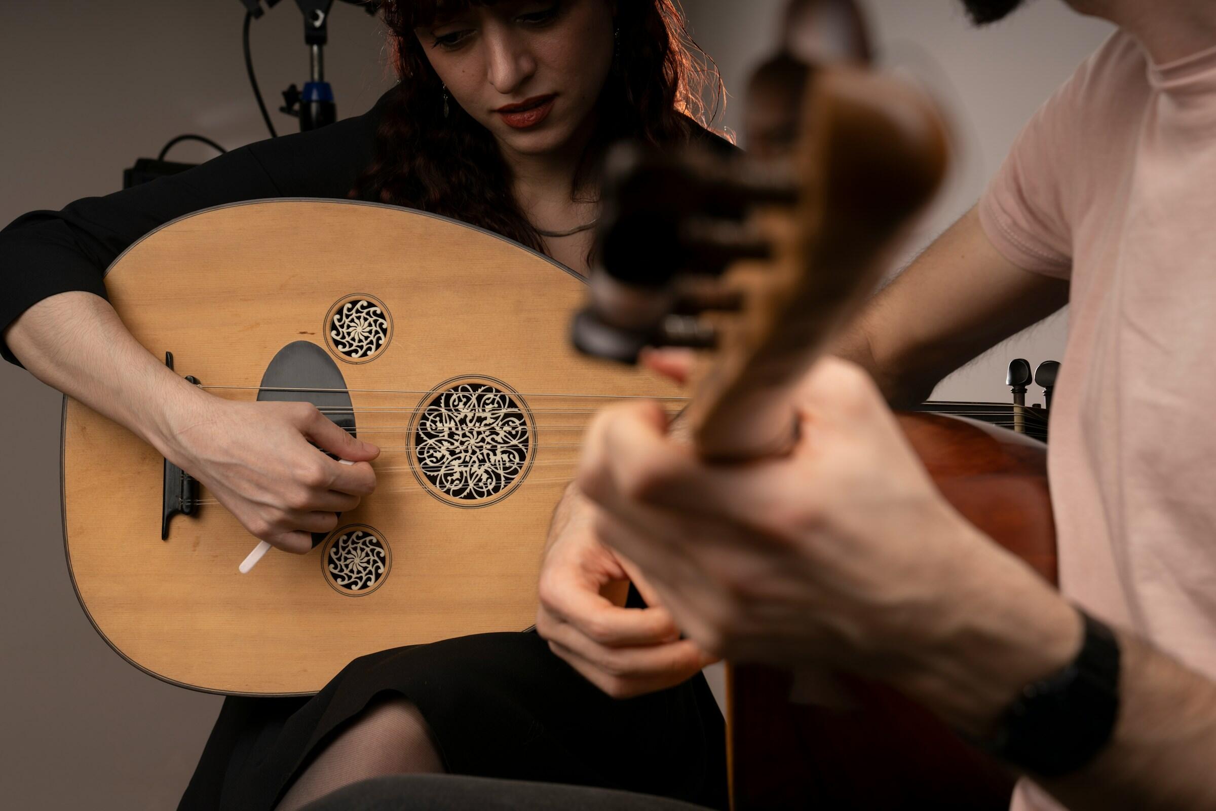 Dos personas tocando instrumentos de cuerda, una el oud y la otra la guitarra, con el foco puesto en sus manos y sus instrumentos.
