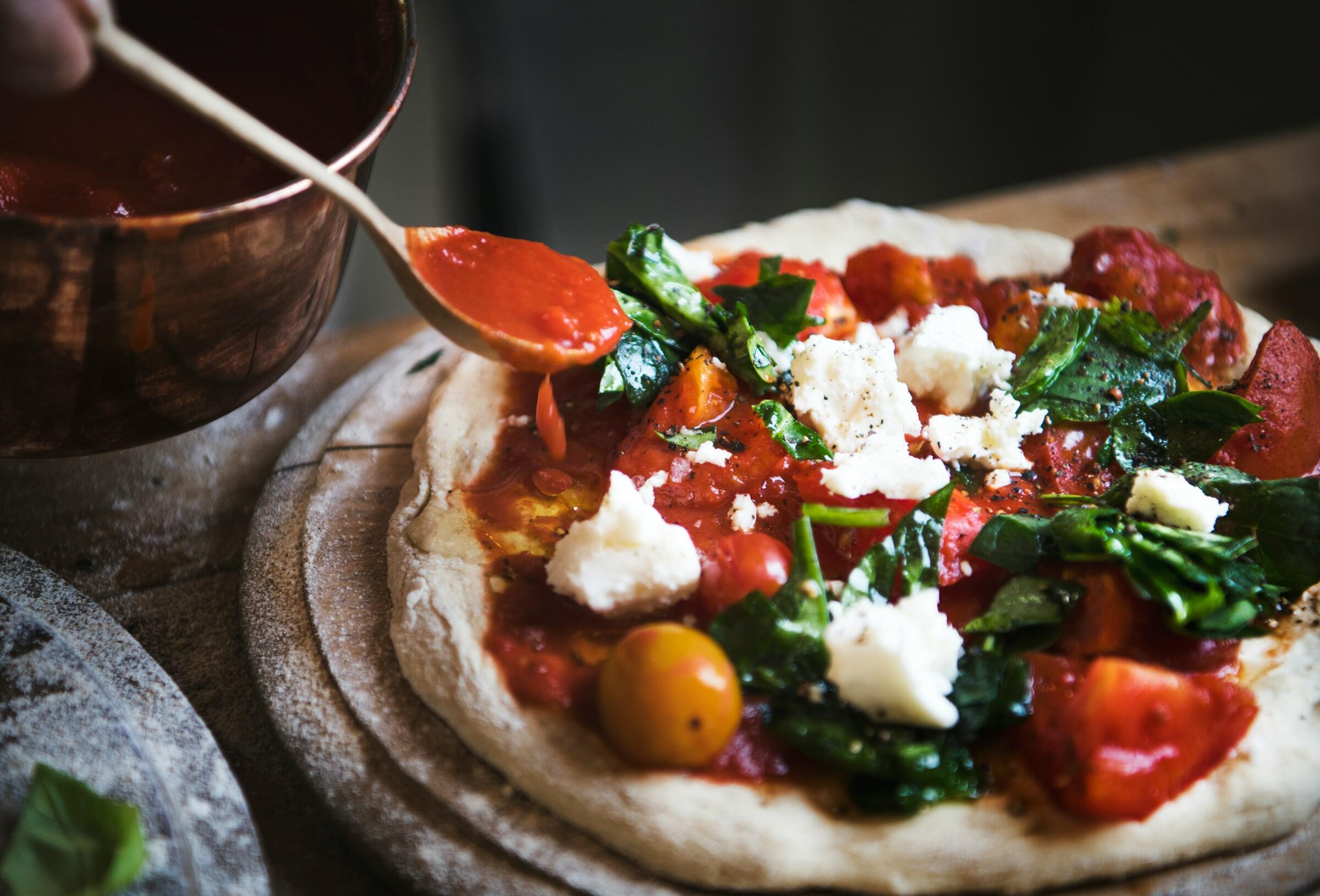 Una cuchara de madera rocía salsa de tomate sobre una pizza cruda cubierta con espinacas, tomates y trozos de queso en una tabla de madera.