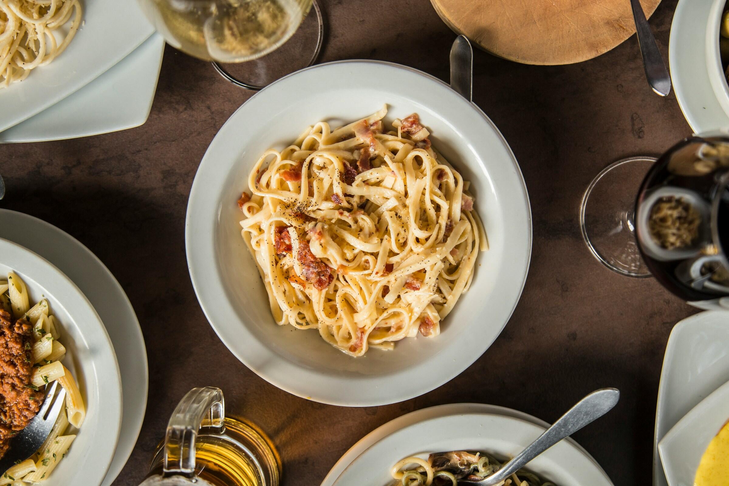 Un plato de fettuccine cremoso con trocitos de bacon, rodeado de platos de otros platos italianos y copas de vino.
