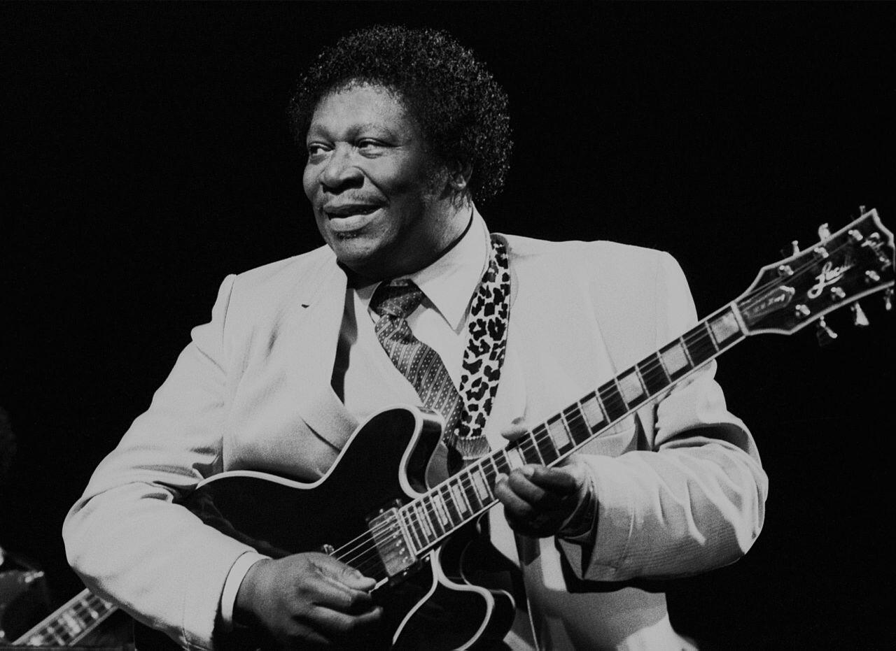 el músico bb king vestido con un traje claro toca la guitarra eléctrica en el escenario, rodeado por un fondo oscuro, lo que evoca una atmósfera de actuación enérgica.