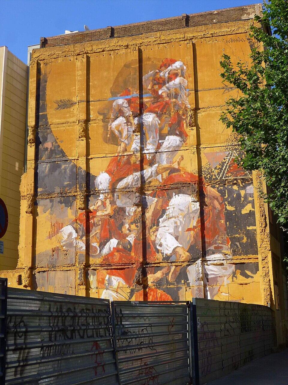 Un vibrante mural en la fachada de un edificio representa figuras en poses dinámicas sobre un fondo amarillo, enmarcadas por el entorno urbano.