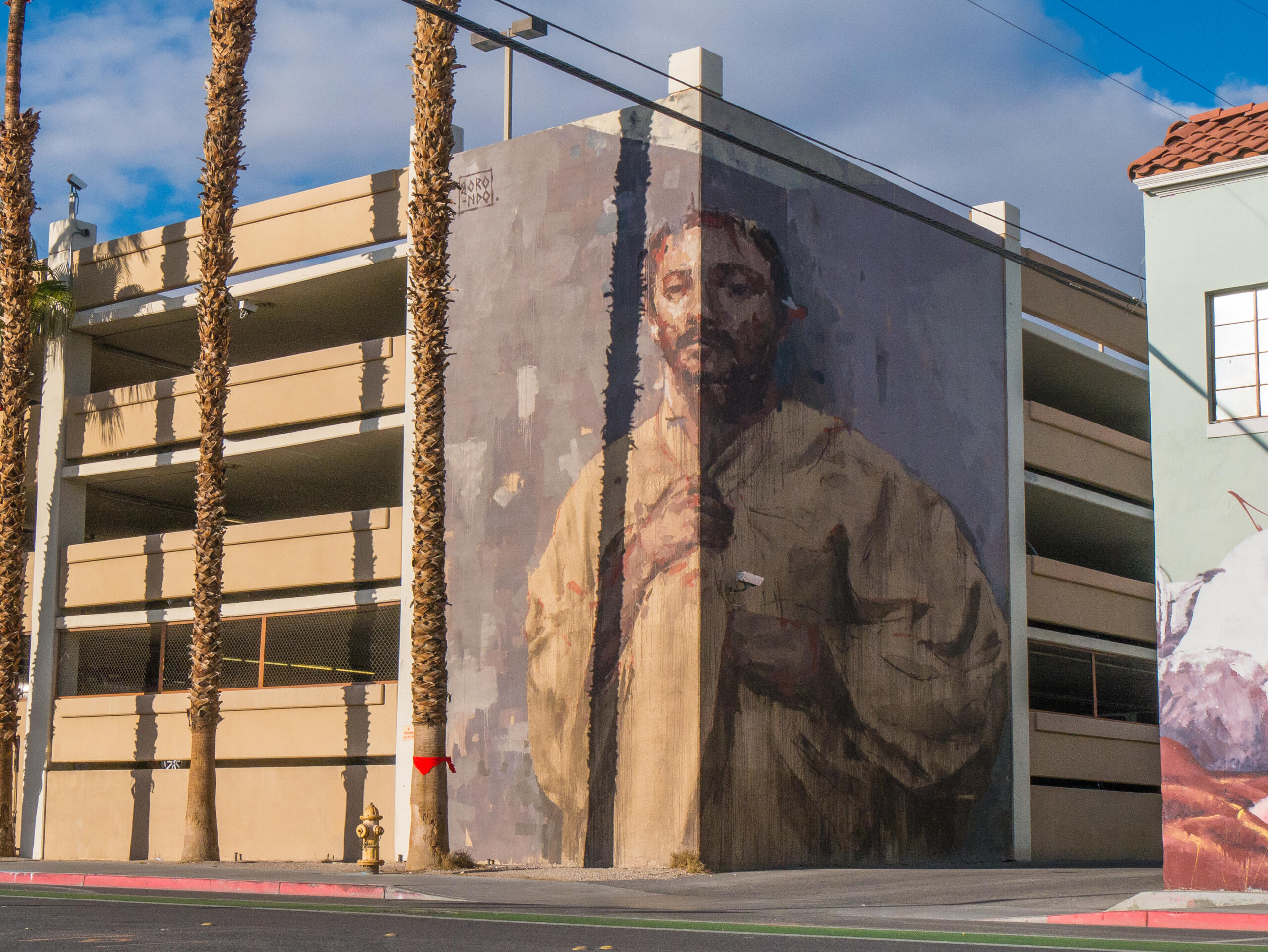 La imagen muestra un gran mural urbano pintado en la fachada lateral de un edificio, representando el retrato de un hombre con un estilo expresivo y pictórico.La obra está integrada en un entorno urbano con palmeras, cables y un aparcamiento, lo que refuerza su carácter contemporáneo.Los tonos apagados y las pinceladas visibles transmiten una sensación de introspección y fuerza emocional.