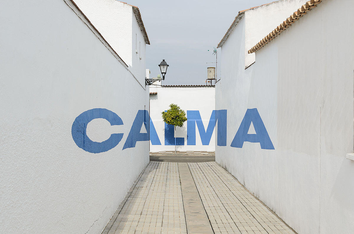 Un callejón estrecho, con paredes blancas, muestra la palabra «CALMA» en grandes letras azules y un pequeño árbol al final, creando una atmósfera serena.