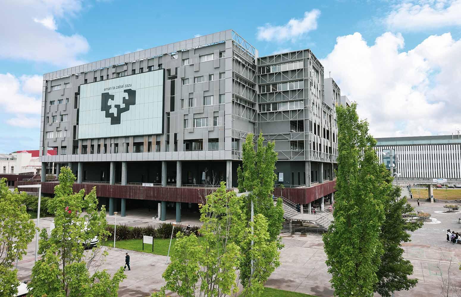 Edificio universitario moderno con un gran logotipo geométrico en la fachada, rodeado de frondosos árboles. El cielo es de un azul intenso con algunas nubes dispersas.