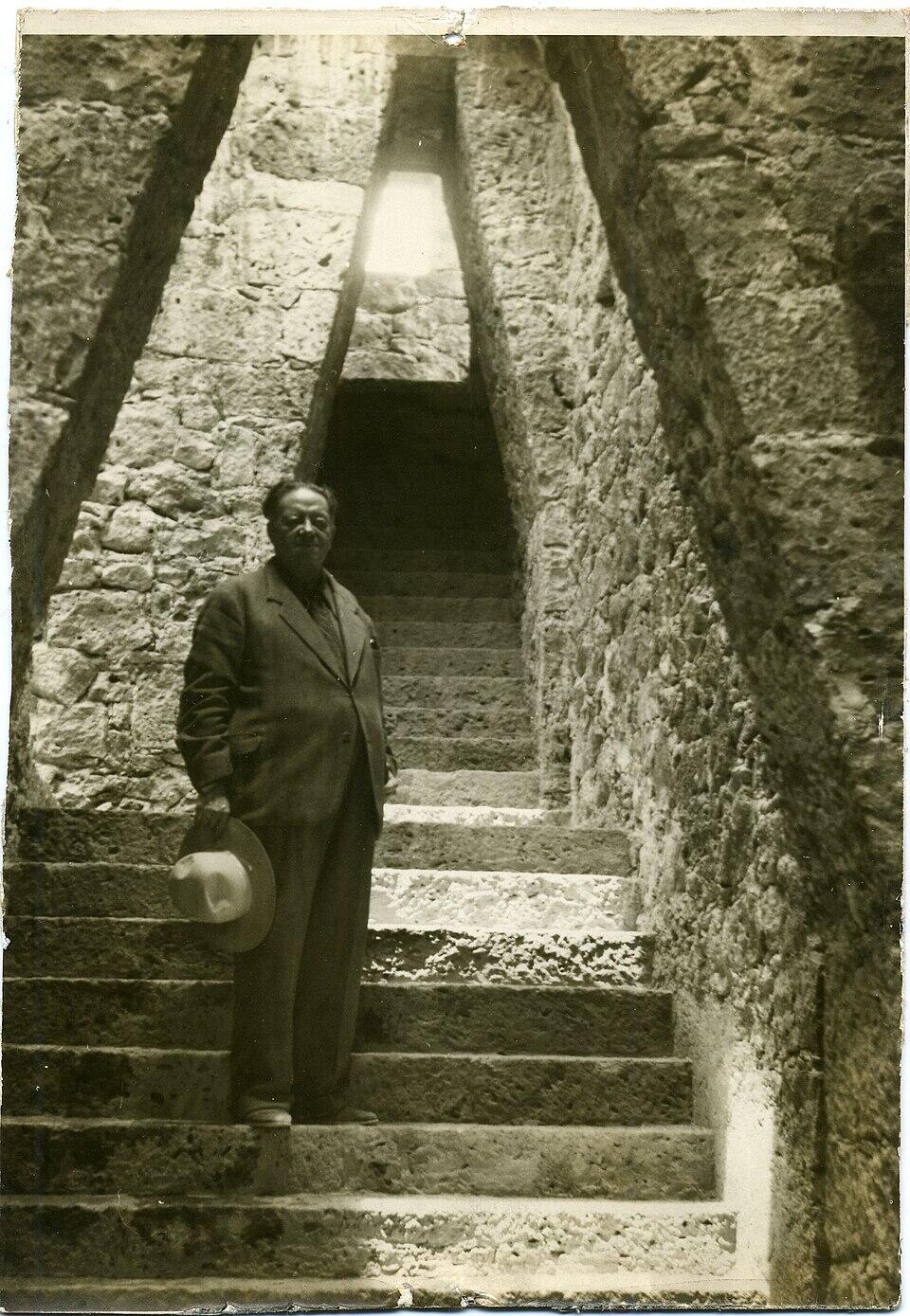 Diego rivera con traje y sombrero está de pie sobre unos escalones de piedra, rodeado por una pared de piedra texturizada, que conduce a un pasillo poco iluminado en la parte superior.