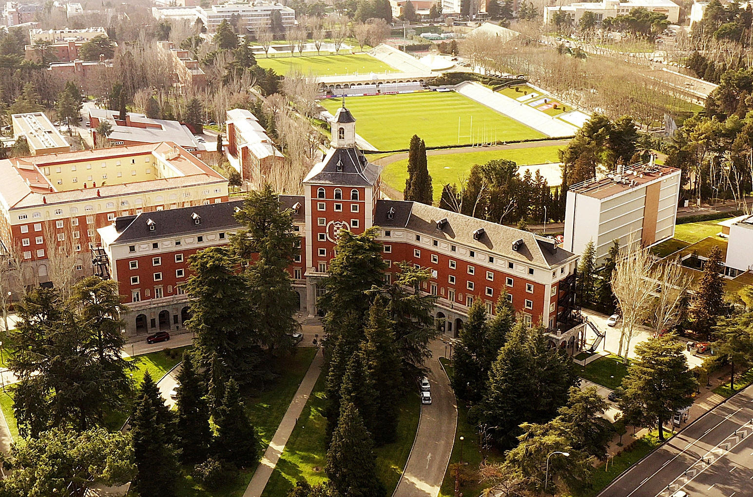 Vista aérea de un campus universitario en la que destaca un edificio de ladrillo rojo con una torre central, rodeado de frondosos árboles y senderos. Al fondo se divisan campos deportivos verdes y otros edificios del campus, lo que crea un ambiente académico y sereno.