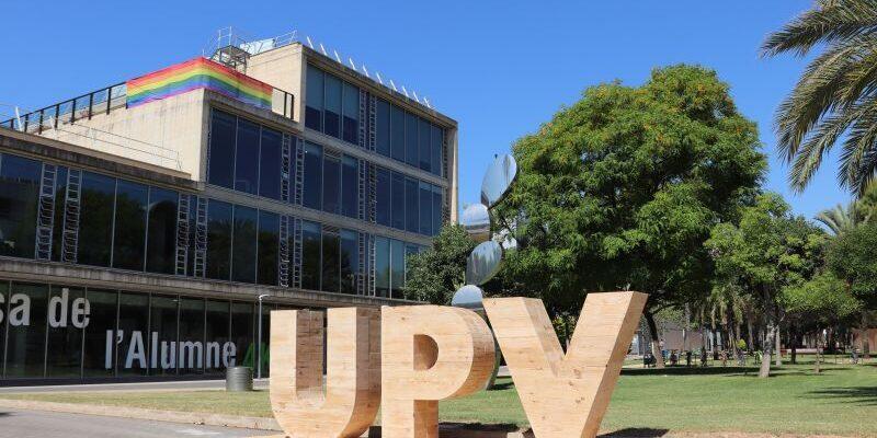 Las letras de madera «UPV» se alzan frente a un moderno edificio acristalado adornado con una bandera arcoíris. Los árboles verdes y el cielo azul claro crean una escena llena de vida y acogedora.