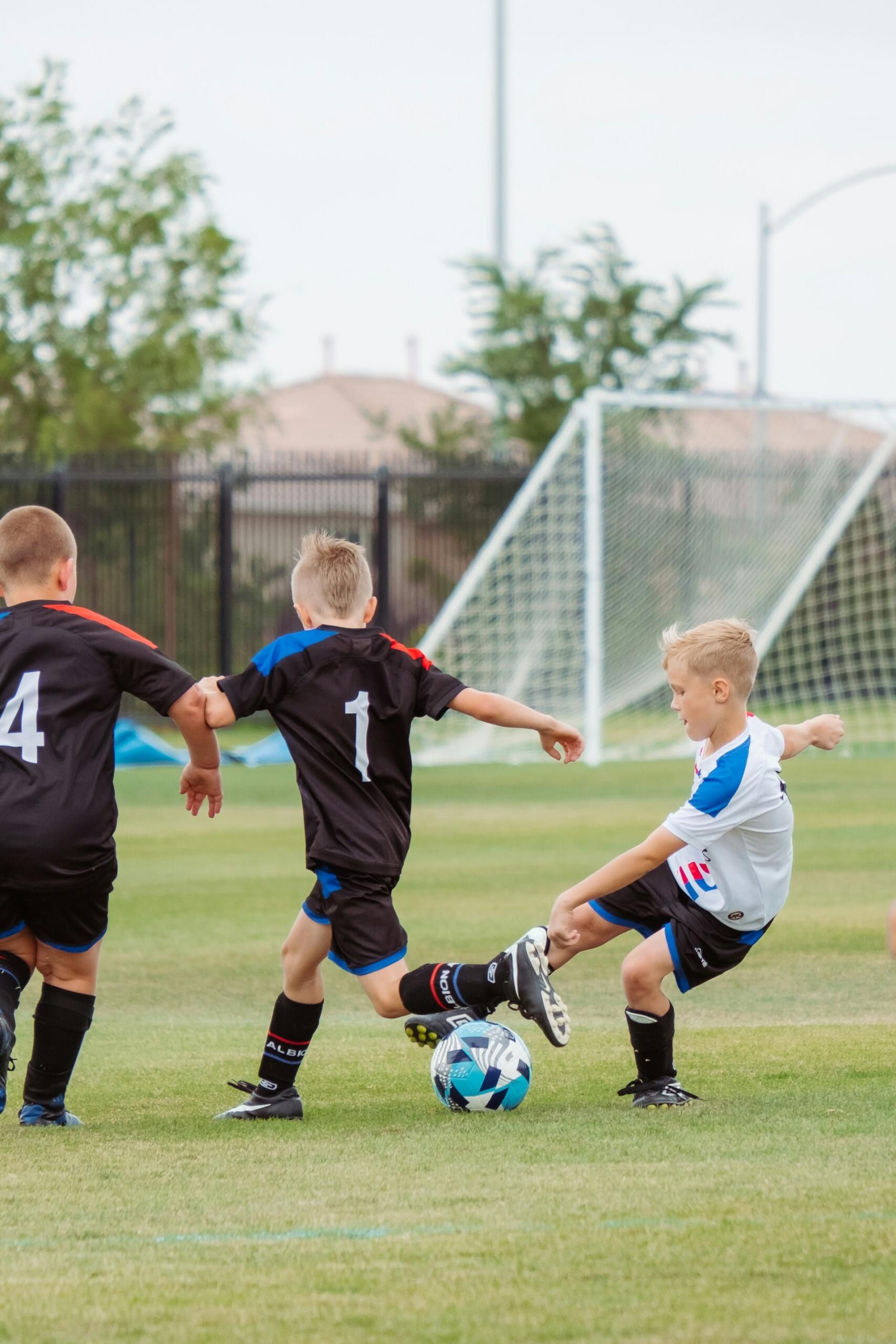 Tres niños pequeños juegan al fútbol en un campo de césped: dos llevan camisetas negras y uno blanca. Corren con energía tras el balón cerca de la portería.