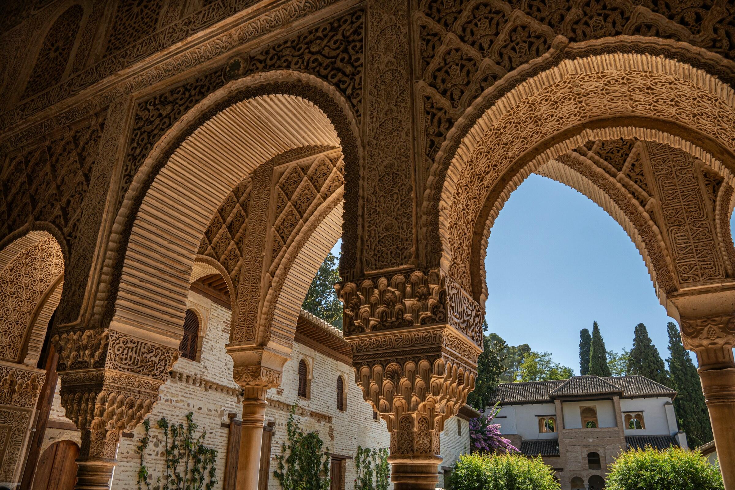 Arcos tallados con gran detalle enmarcan un tranquilo patio de la Alhambra, resaltando los intrincados detalles arquitectónicos islámicos y la exuberante vegetación.