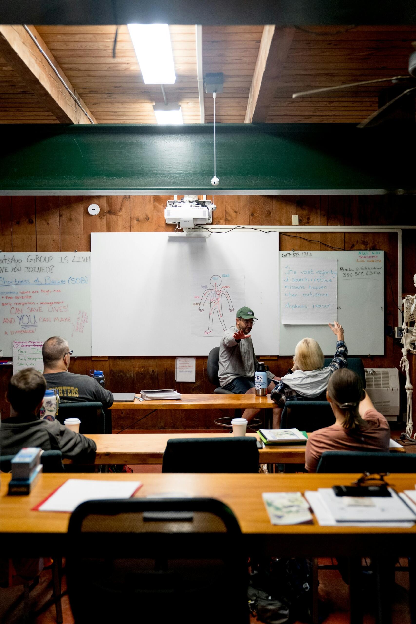 Un aula con cuatro personas sentadas en sus pupitres; un profesor señala con un gesto un diagrama de un esqueleto en la pizarra. La sala tiene un ambiente acogedor y rústico.