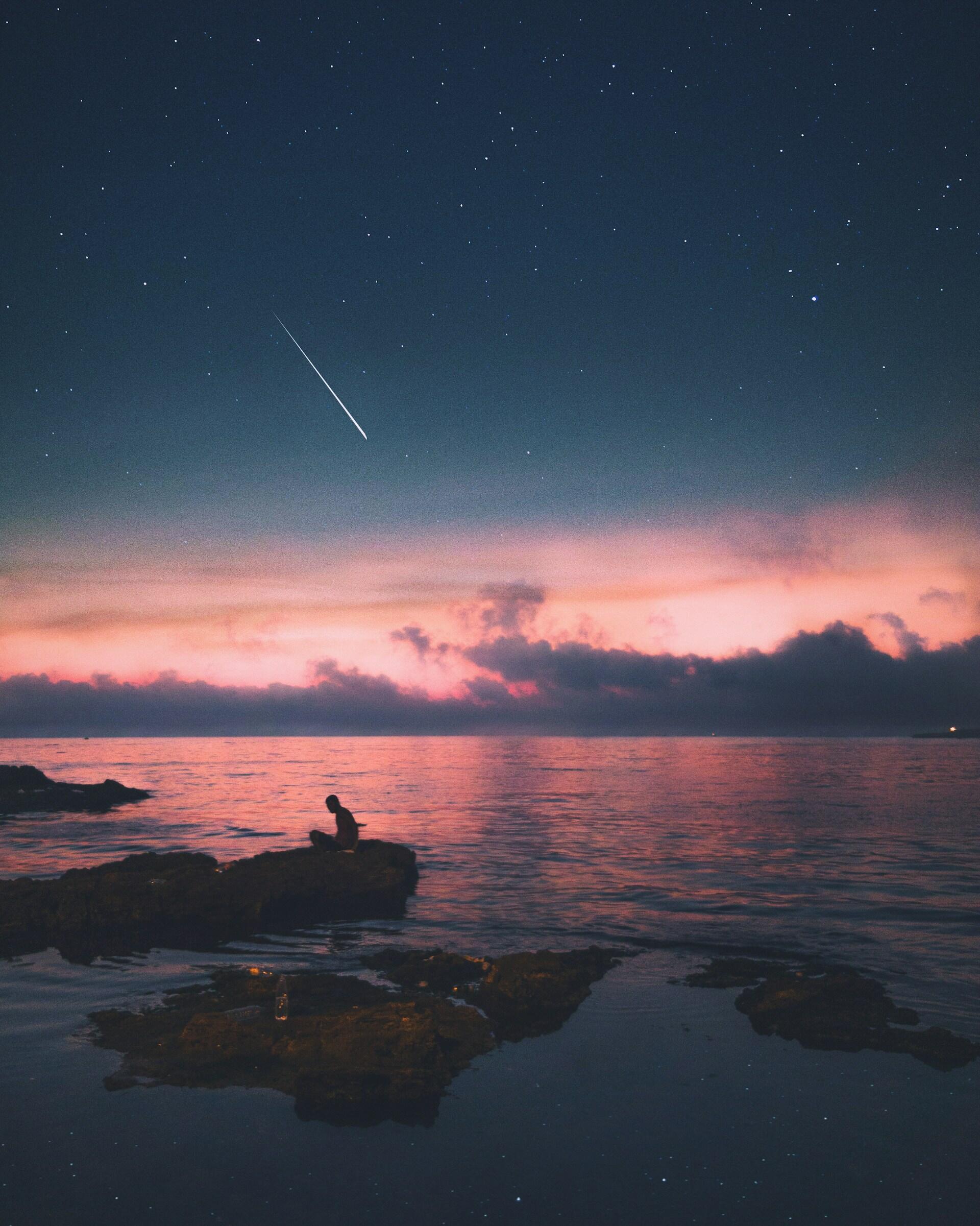 Una persona está sentada sobre una roca junto al mar en una noche estrellada, con una puesta de sol de vivos tonos rosas y morados y una estrella fugaz en el cielo, lo que evoca una atmósfera tranquila.
