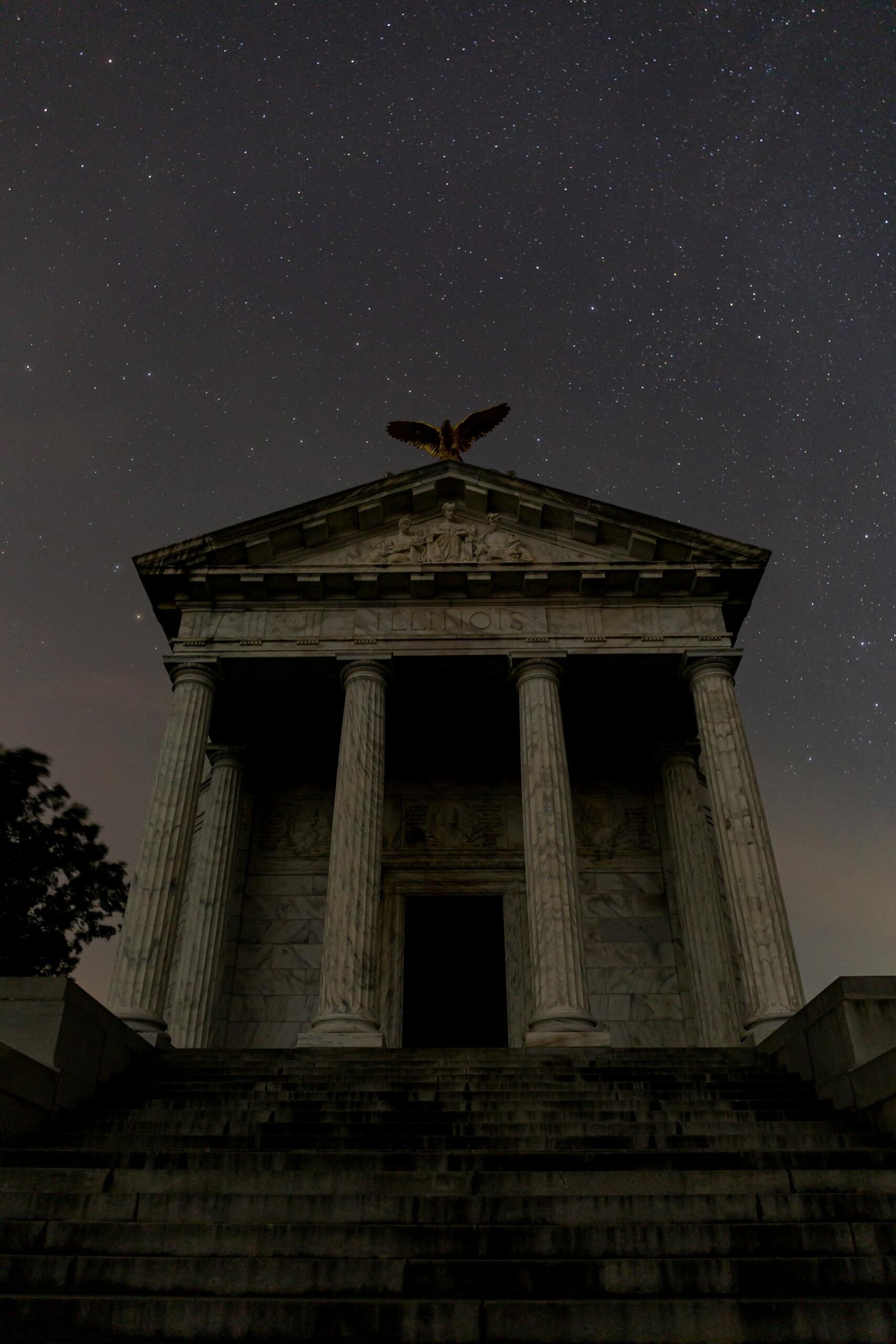 Un edificio antiguo de estilo griego, con una iluminación tenue, altas columnas y una escultura de un águila en lo alto, recortado contra un cielo nocturno estrellado, evoca una atmósfera serena y majestuosa.