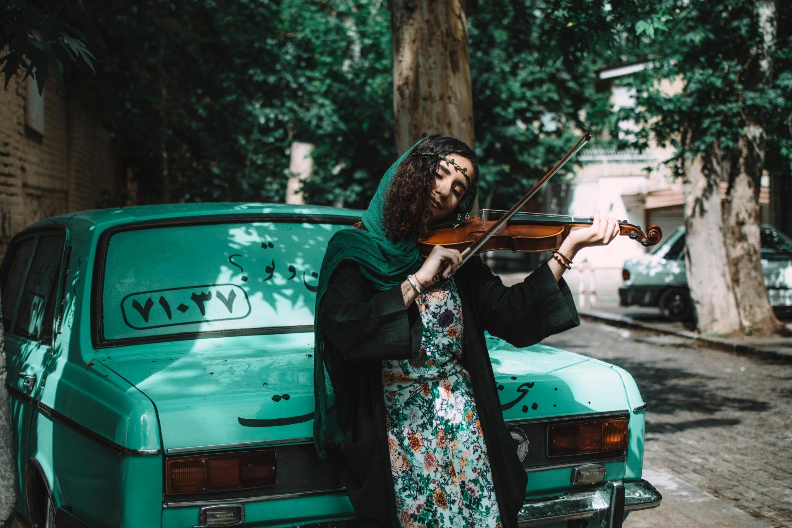 Mujer árabe tocando violín apoyada sobre un auto en la calle.