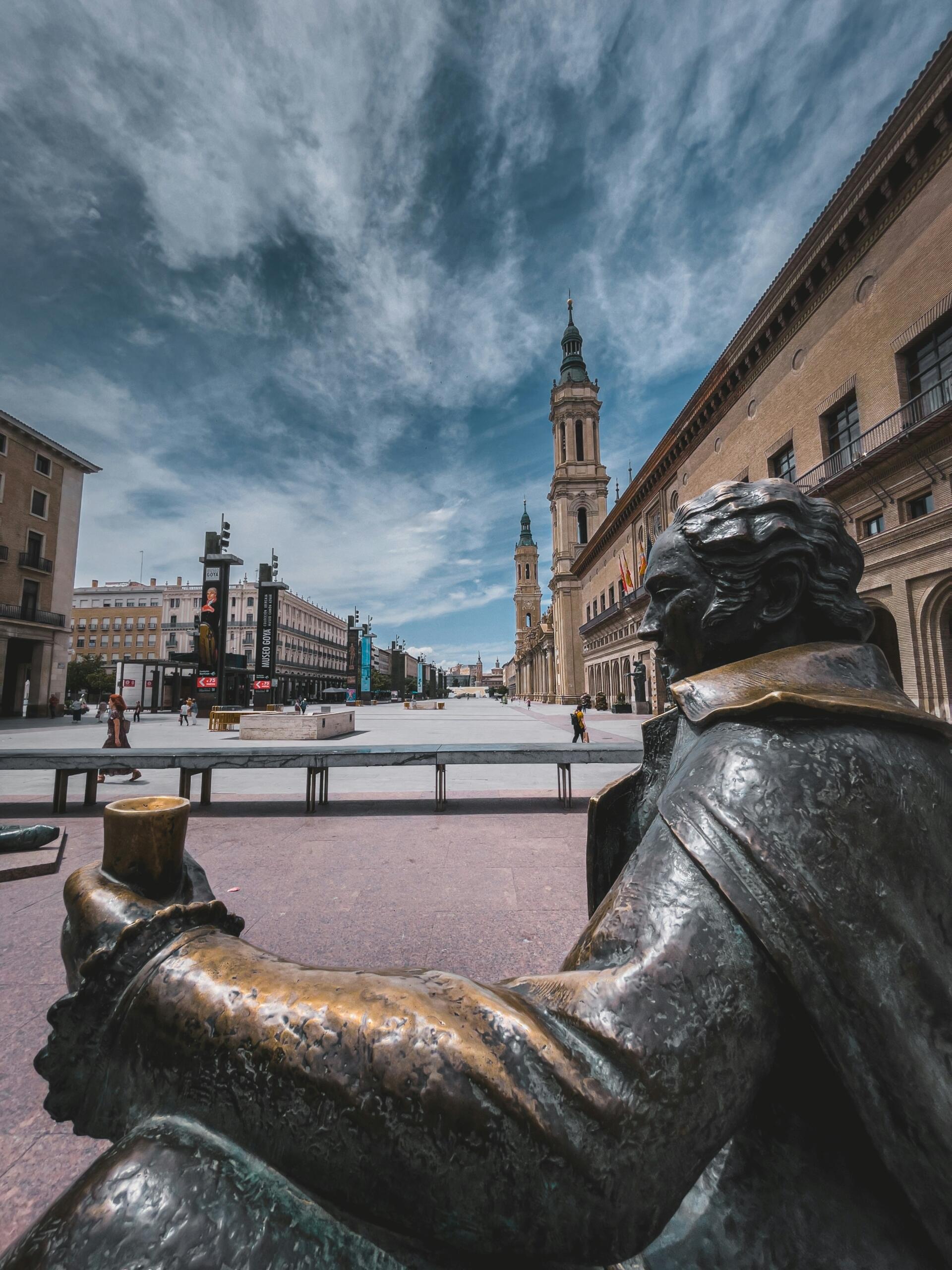 Una estatua de bronce de francisco de golla con la cara levantada domina una amplia plaza, que conduce a una torre y a edificios bajo un cielo azul con nubes dispersas.
