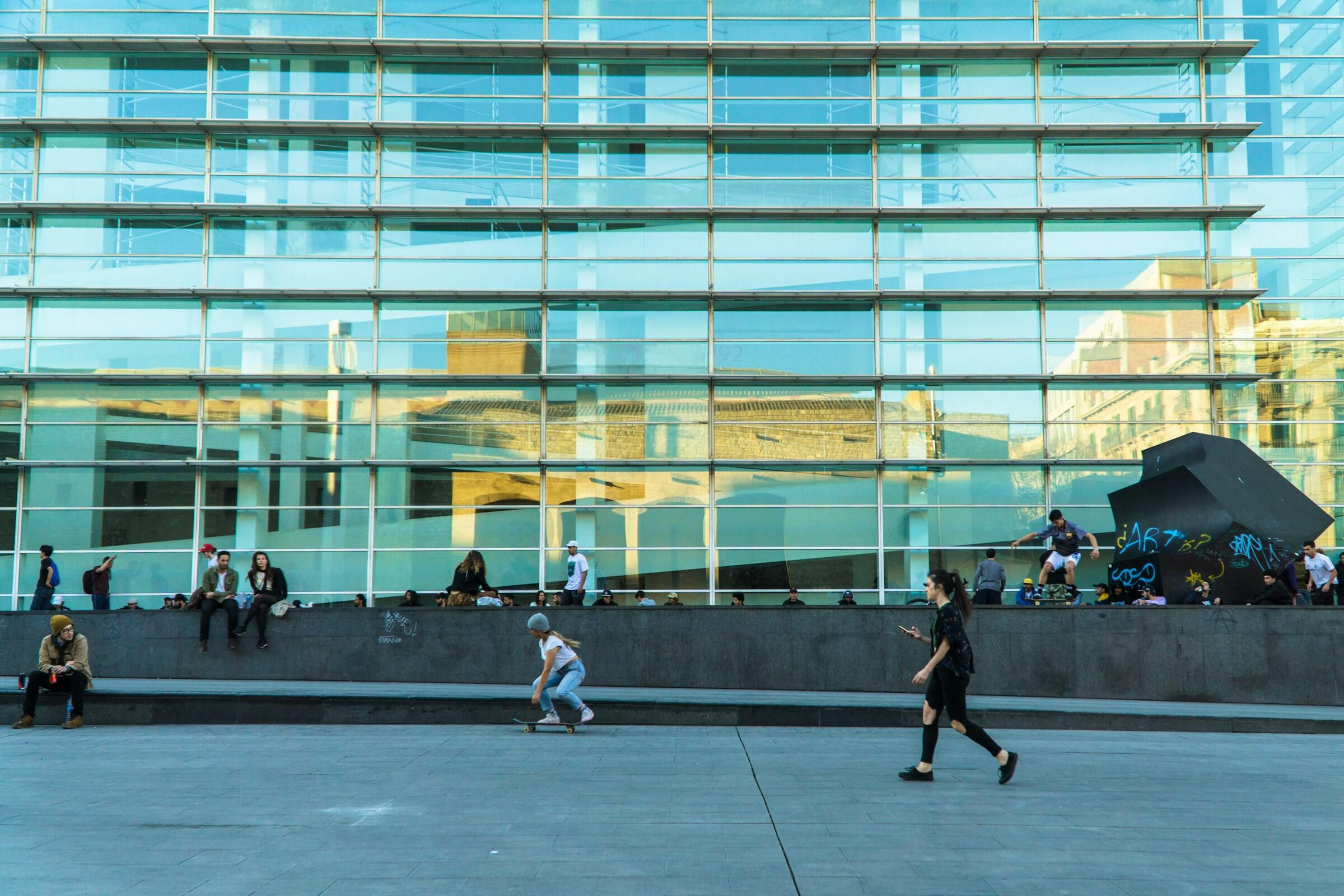 Una bulliciosa plaza urbana muestra a gente sentada y paseando cerca de un moderno edificio de cristal, con reflejos de la arquitectura circundante del edificio del museo de arte contemporaneo de barcelona macba
