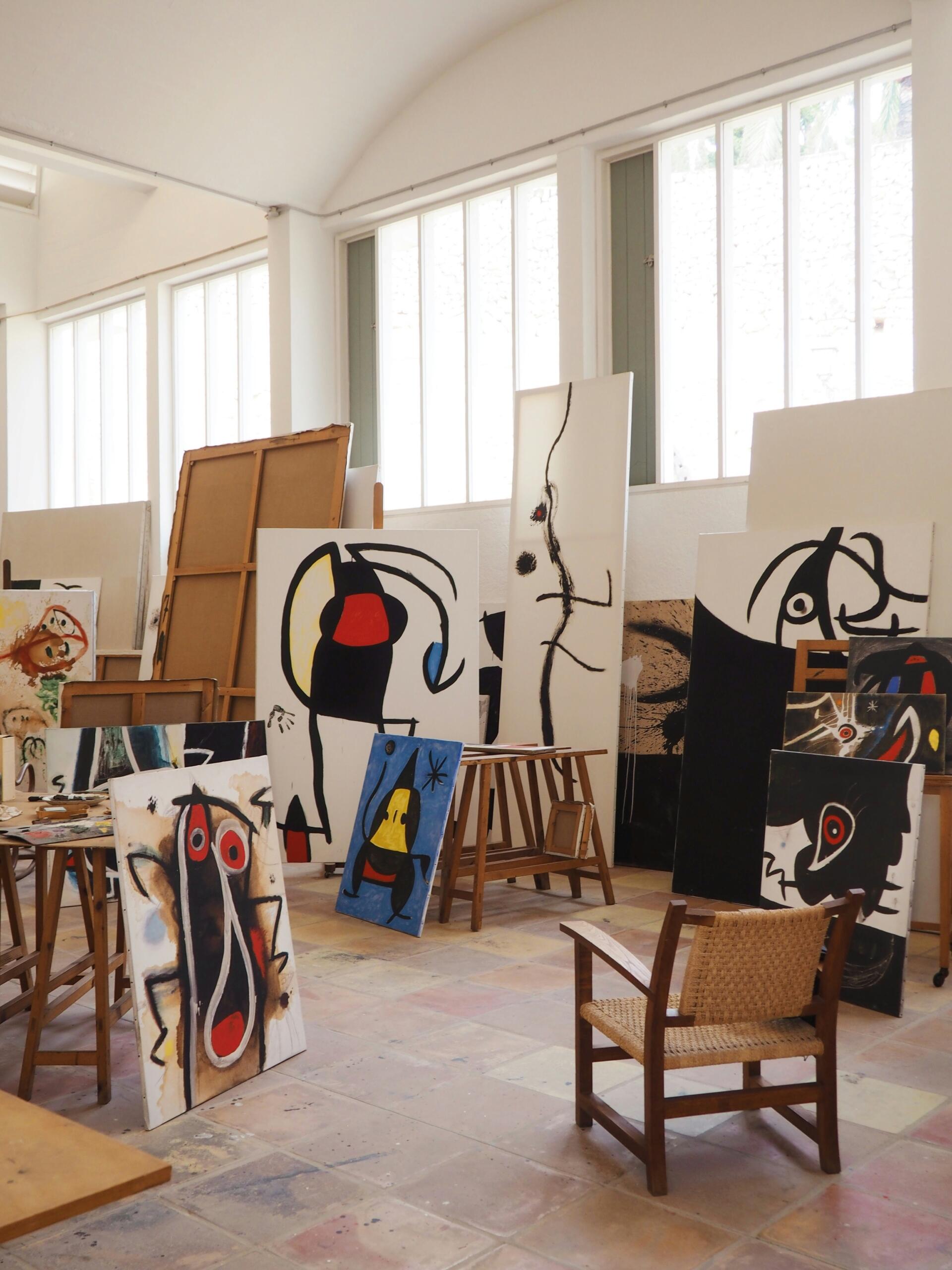 Luminoso estudio de arte lleno de cuadros abstractos de miro sobre caballetes, una silla de madera en primer plano y grandes ventanales que dejan entrar la luz natural.