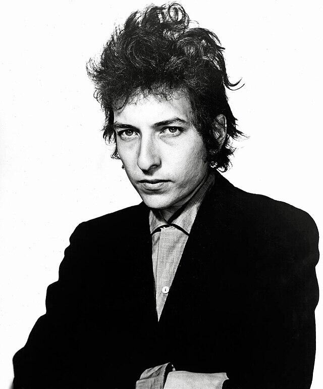 Retrato en blanco y negro de Bob Dylan con el pelo rizado y despeinado llevando una chaqueta negra sobre una camisa con cuello y posando con confianza sobre un fondo blanco.