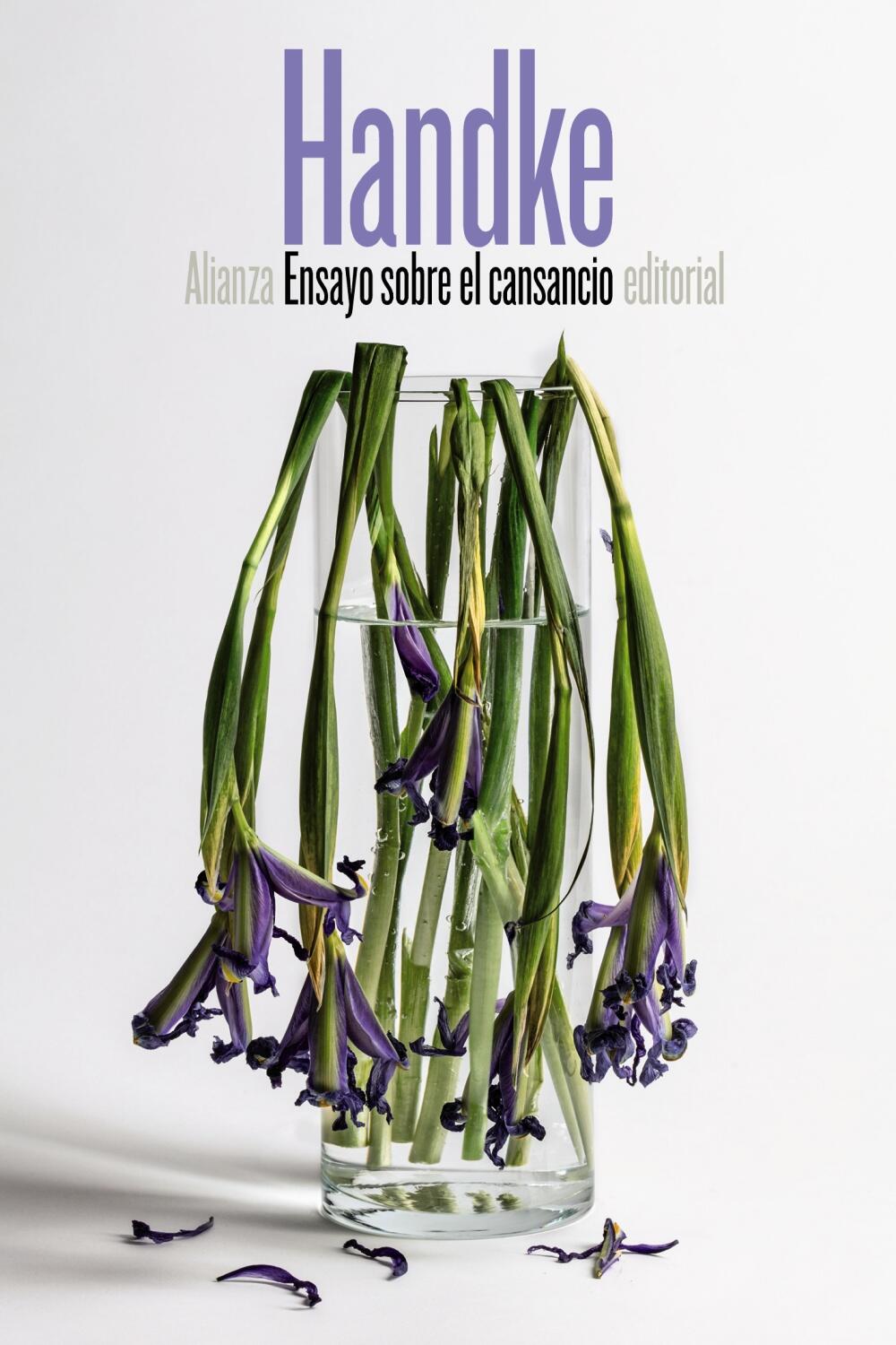Un jarrón de cristal contiene varias flores moradas marchitas con tallos caídos sobre un fondo blanco liso. El texto de arriba dice: Handke. Ensayo sobre el cansancio. Alianza editorial.