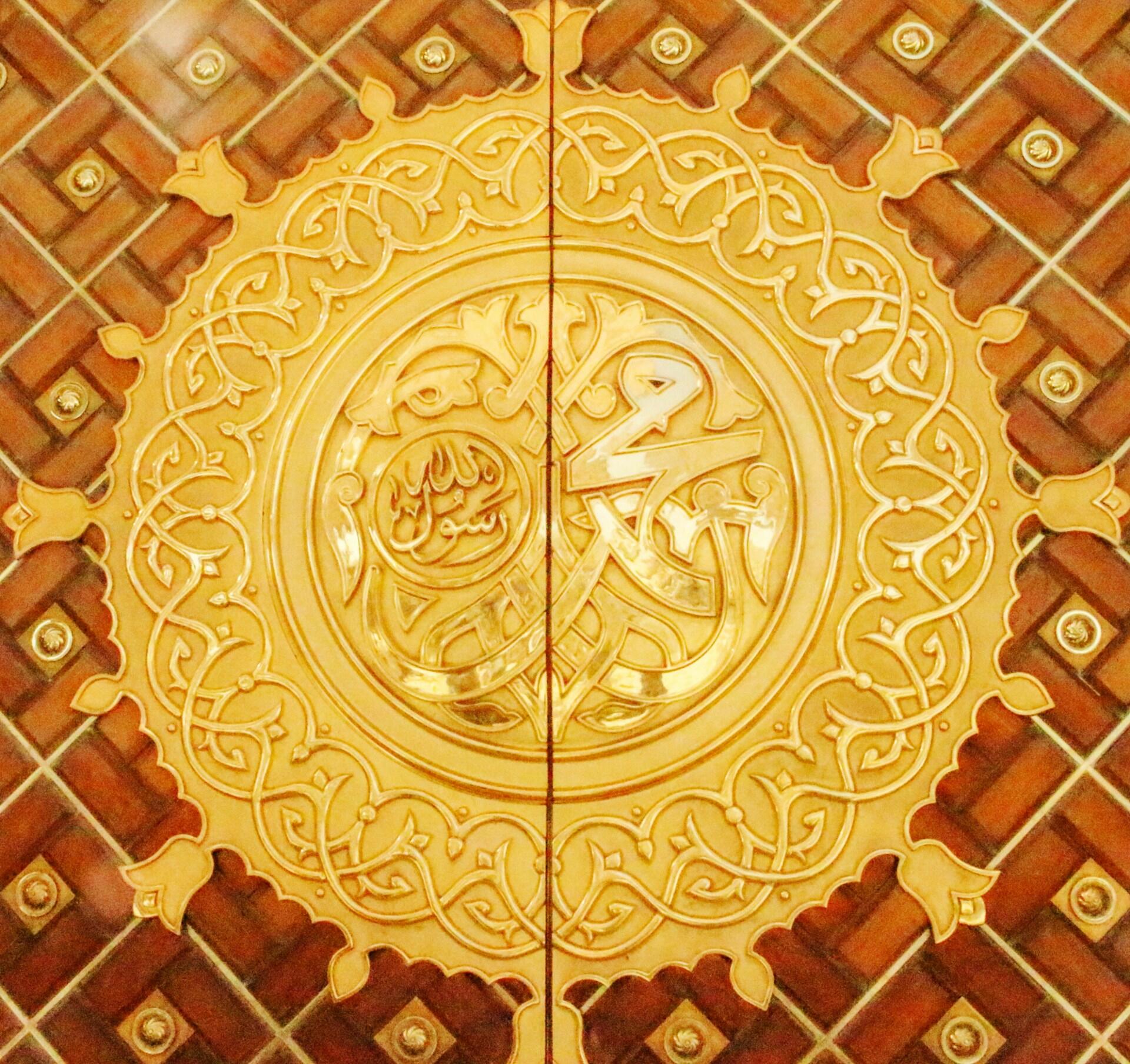 Una ornamentada caligrafía árabe dorada llena un medallón circular, enmarcado por volutas florales y azulejos geométricos, lo que crea un ambiente cálido y solemne.