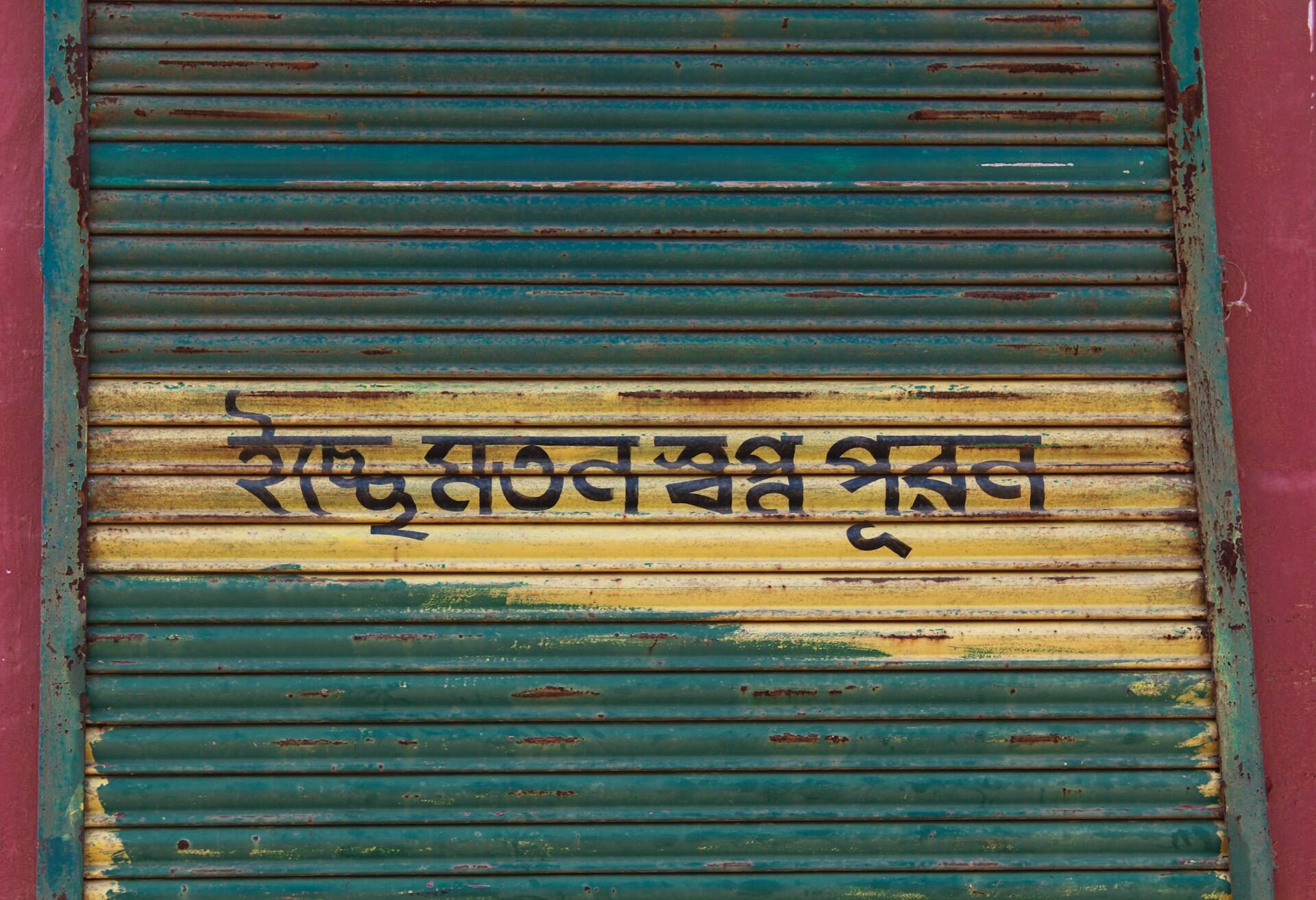 Persiana ondulada de color verde azulado y amarillo, desgastada por el paso del tiempo y cubierta de grafitis en devanagari oscuro, lo que crea un ambiente urbano, envejecido y descarnado.