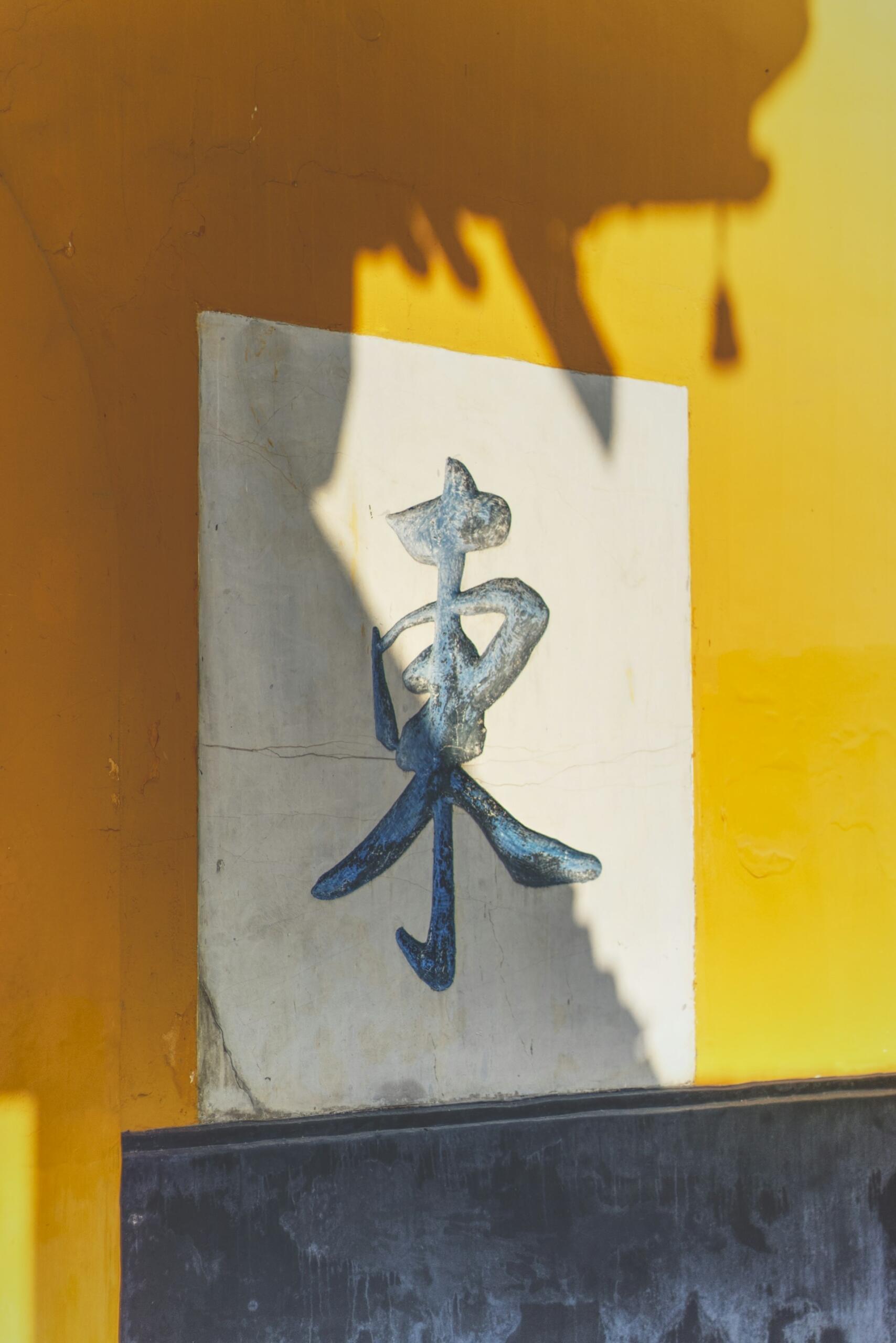 Una pared amarilla iluminada por el sol con un cartel blanco desgastado en el que se ve una figura abstracta azul que baila; las sombras marcadas crean una escena callejera cálida y misteriosa.