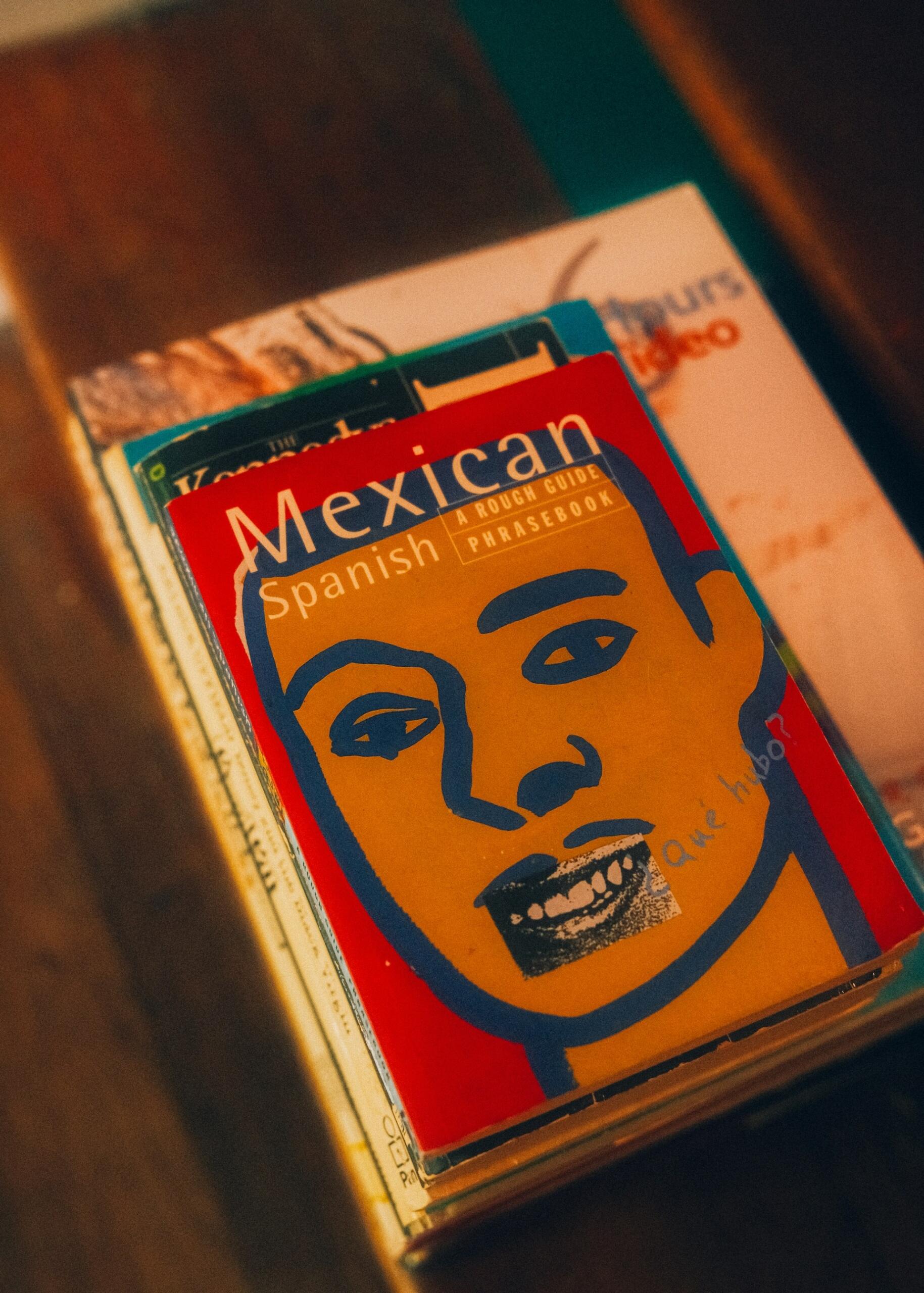 Sobre unos libros antiguos descansa un colorido manual de frases en español mexicano, cuya portada roja, naranja y azul luce una llamativa ilustración de un rostro.