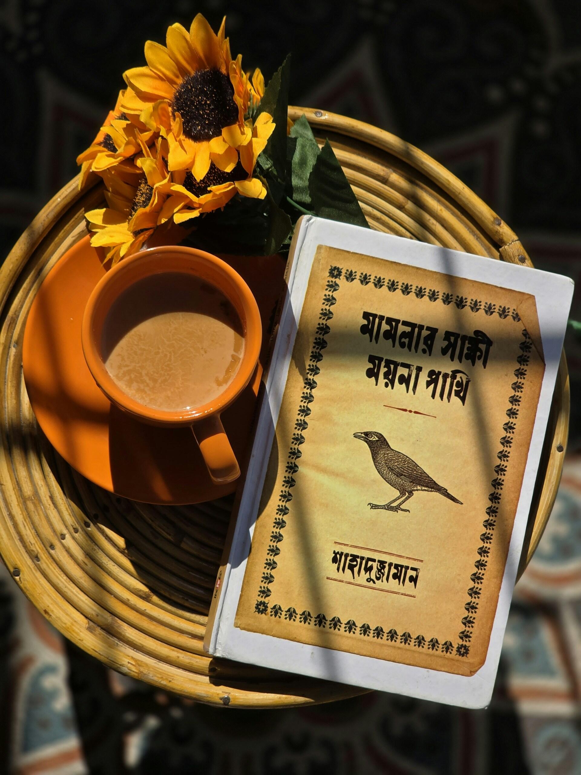 Vista aérea de una bandeja tejida con un girasol, té humeante en una taza naranja y un libro escrito en devanagari con la ilustración de un pájaro, todo ello bañado por una cálida luz.