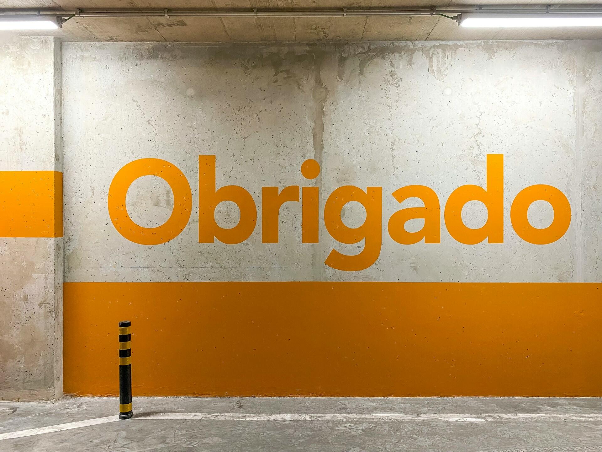 La palabra «Obrigado» (que en portugués significa «gracias») pintada en naranja sobre la pared desgastada de un aparcamiento de hormigón, creando un mensaje llamativo y acogedor.