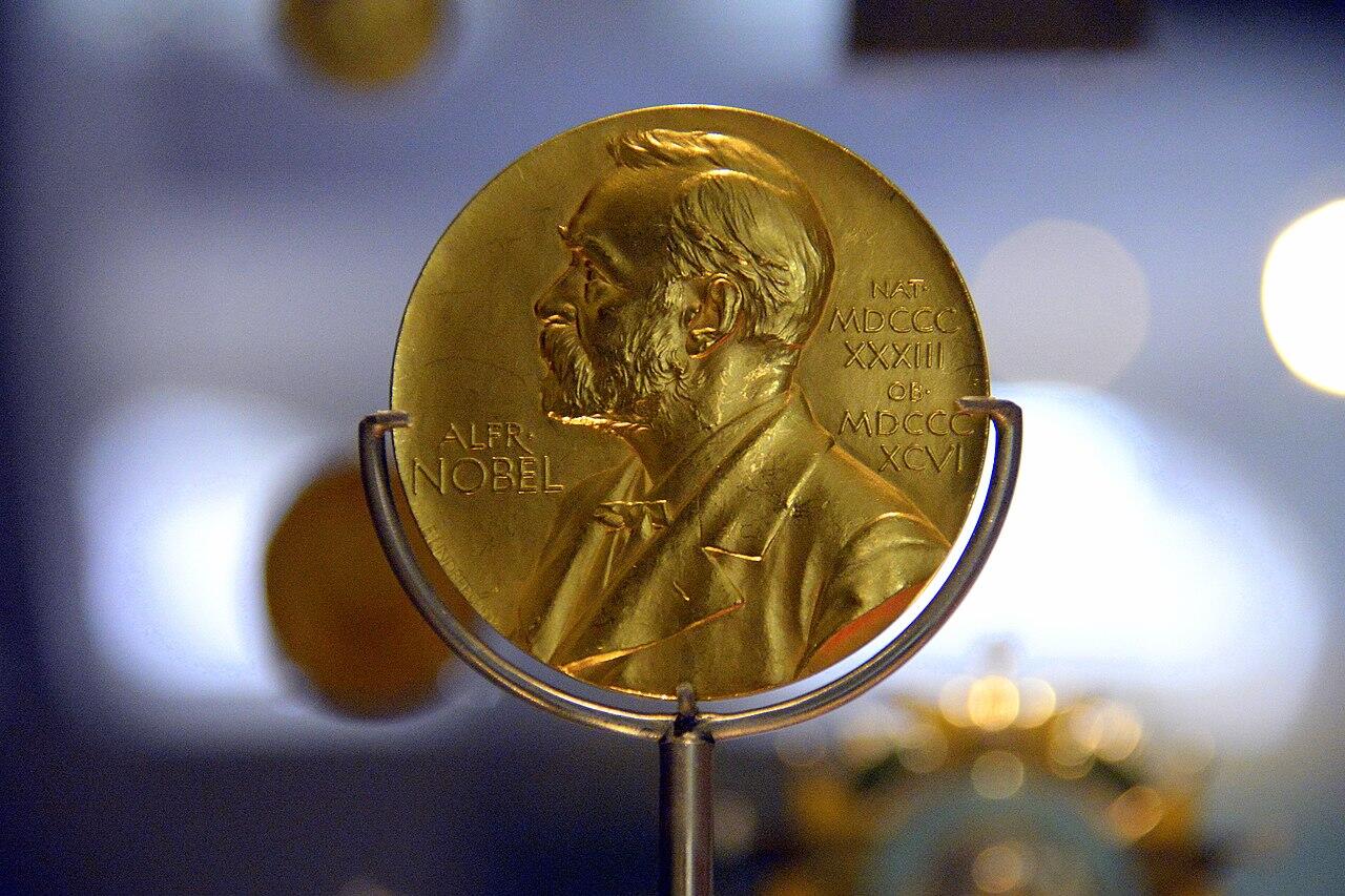 Una medalla de oro del Premio Nobel con el perfil de Alfred Nobel y unas inscripciones se muestra sobre un fondo difuminado, lo que transmite prestigio.