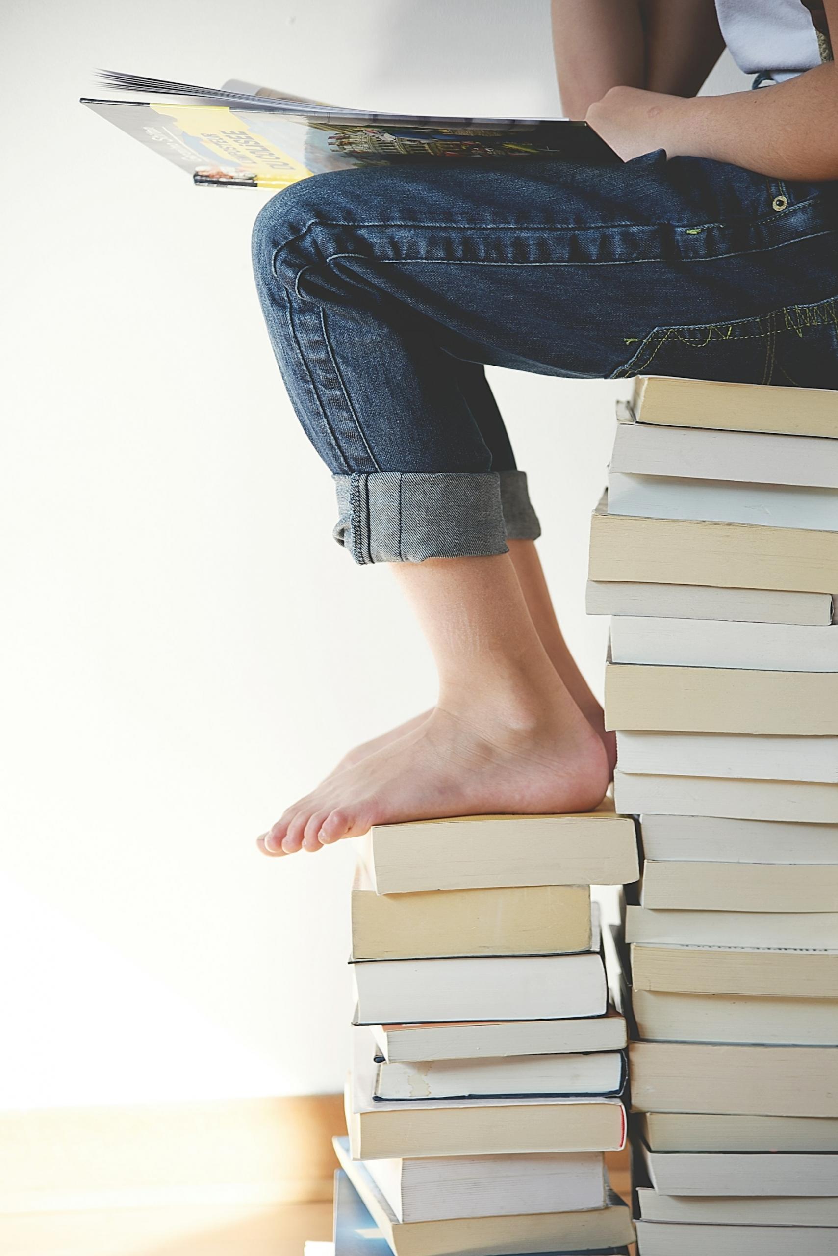 una persona sentada sobre un montón de libros y con sus pies también sobre otra ruma de libros.
