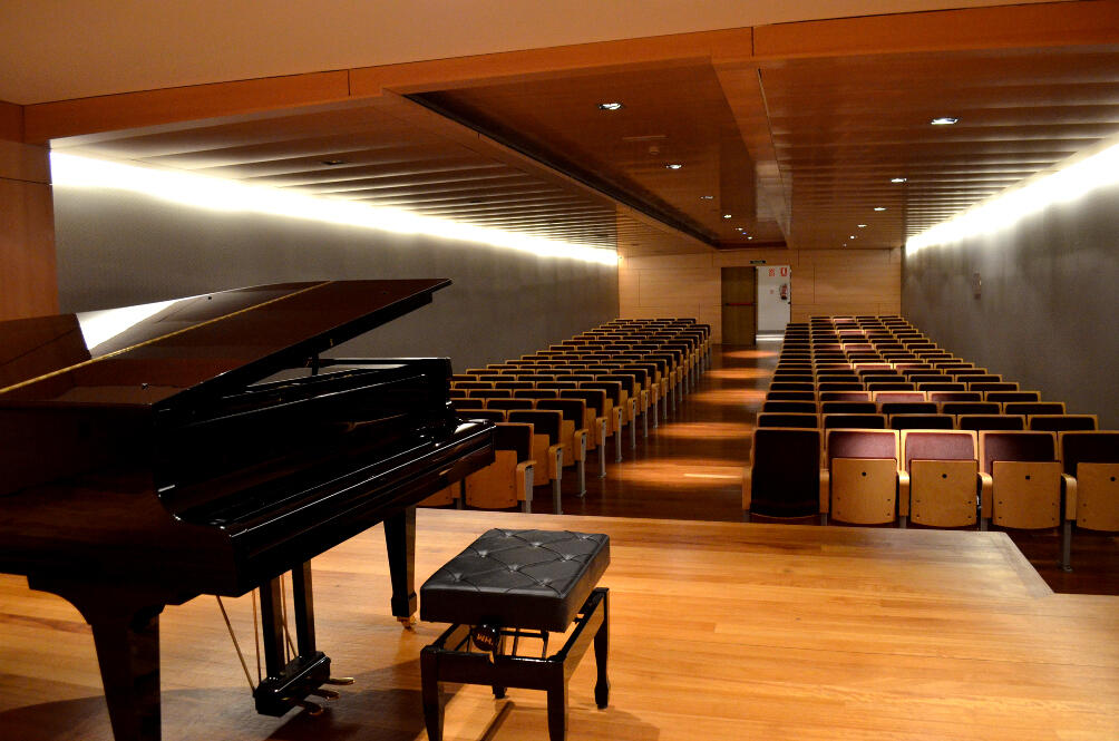 Un piano de cola y un banco en primer plano, con vistas a un moderno auditorio lleno de filas de sillas de madera.
