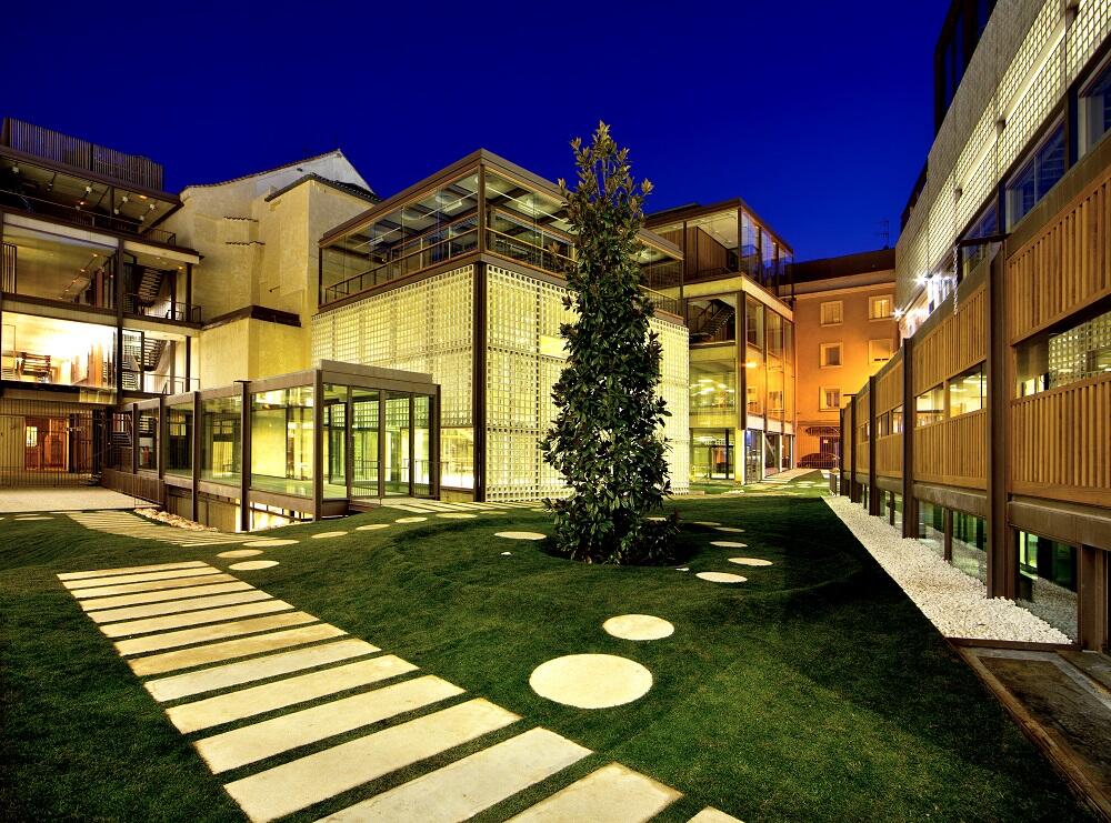 Patio moderno por la noche con edificios acristalados, césped verde, piedras para cruzar el agua y un árbol iluminado en medio de la arquitectura contemporánea.