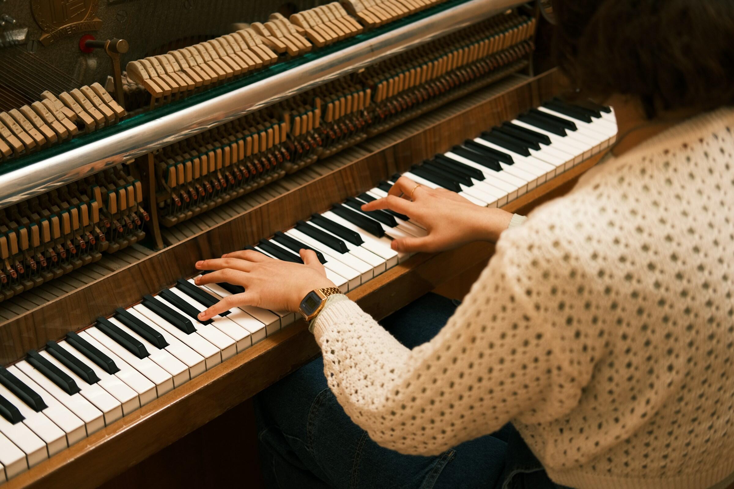 Una persona toca el piano, con las manos sobre las teclas, vestida con un jersey blanco de punto y un reloj, y con la mecánica interna del piano a la vista.