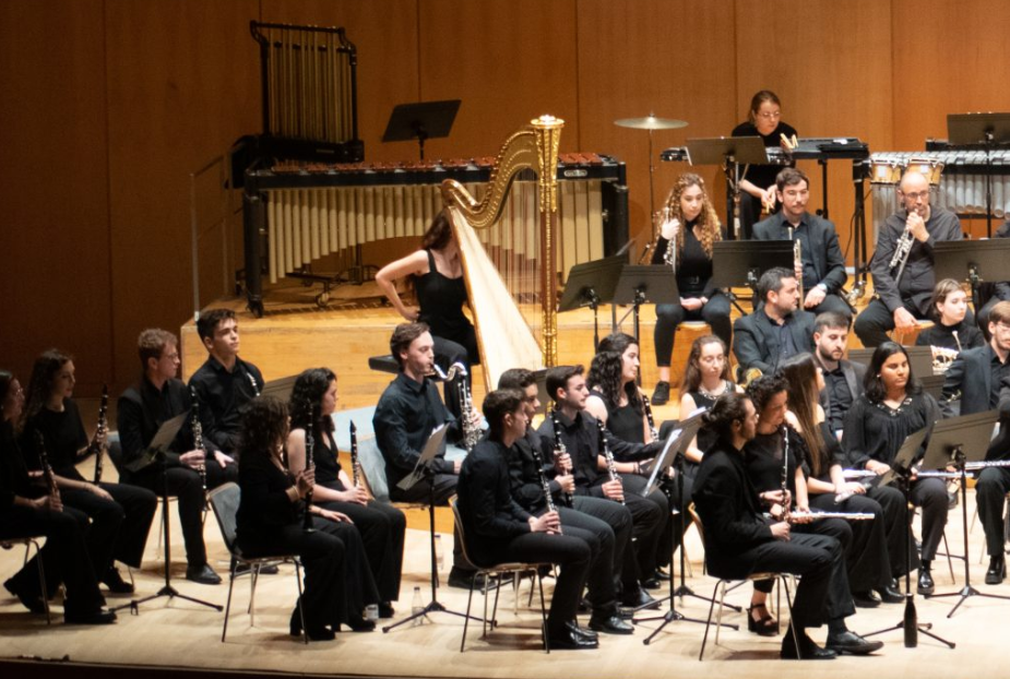Una banda de música actúa en el escenario, con músicos que tocan instrumentos de viento y un arpa, vestidos con trajes negros.