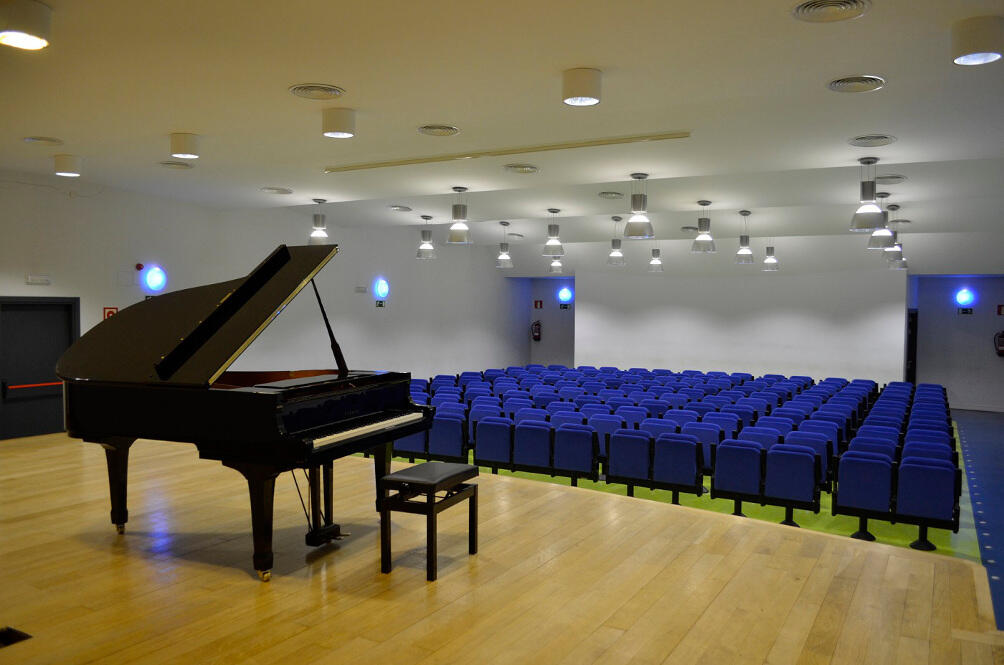 Un piano de cola y un taburete sobre un escenario de madera, frente a un auditorio con filas de sillas azules y una moderna iluminación cenital.