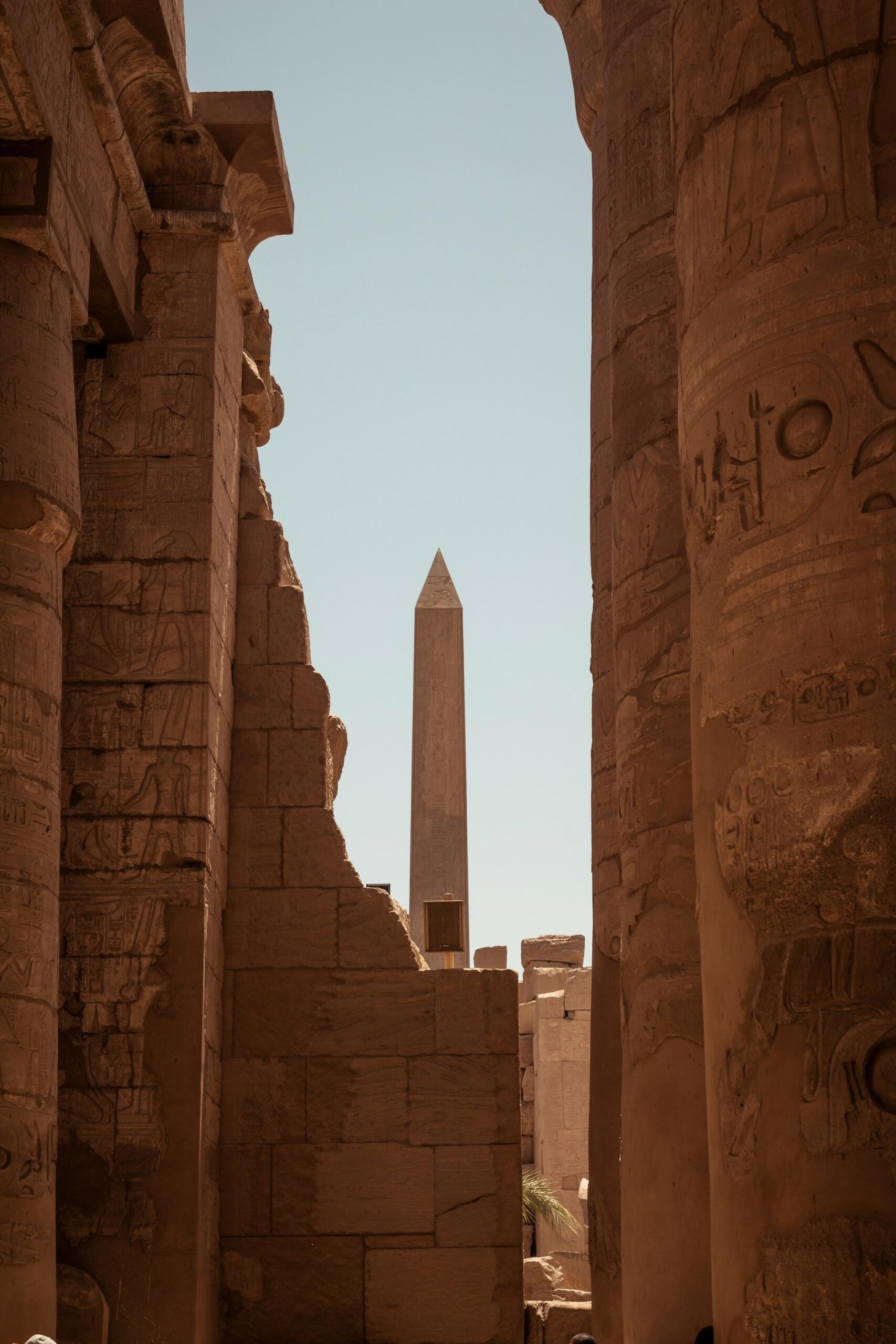 Antiguas columnas de piedra enmarcan un obelisco lejano bajo un cielo despejado. Jeroglíficos y tallas adornan los pilares, evocando una sensación de grandeza histórica.