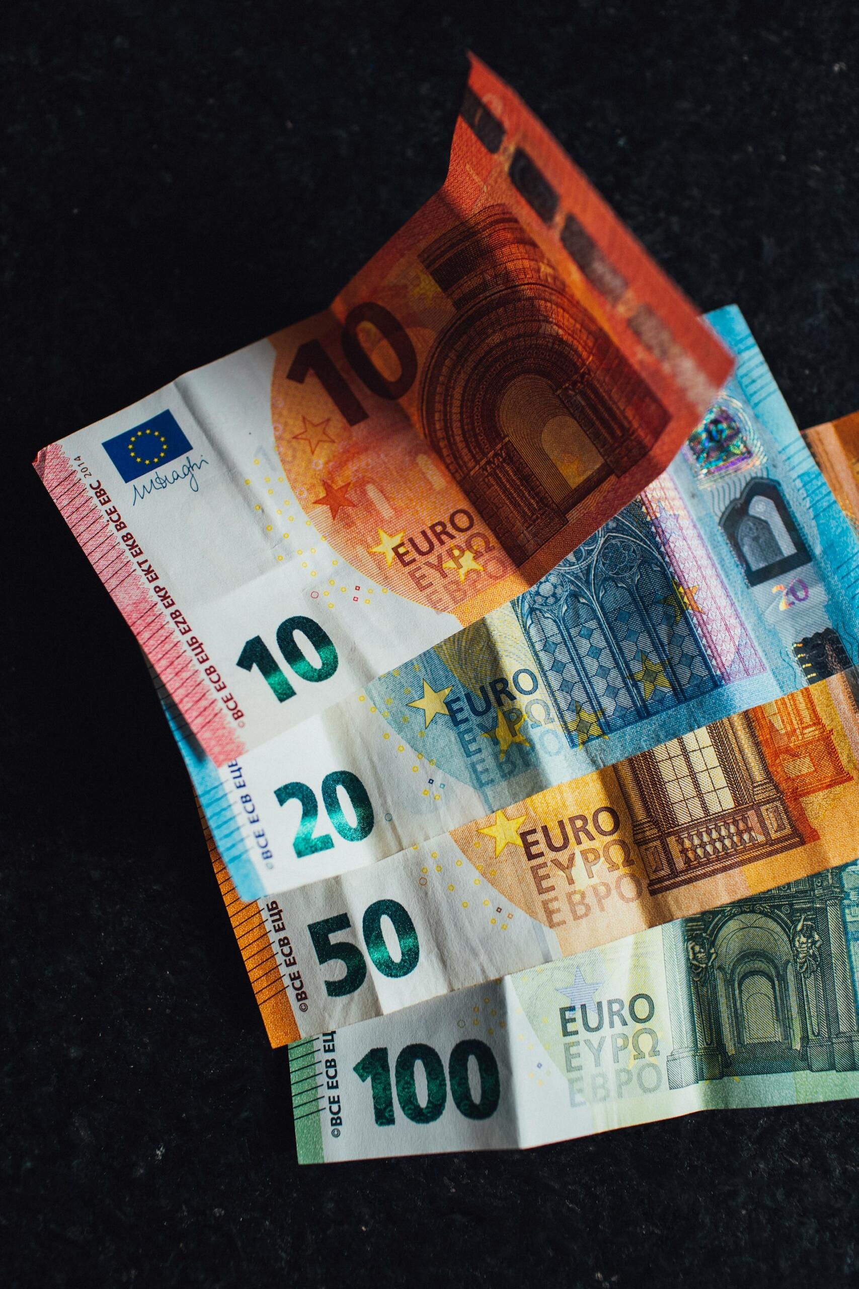 Un colorido abanico de billetes de euro, de 10 a 100 euros, dispuestos sobre una superficie oscura. Cada billete presenta distintos diseños arquitectónicos.