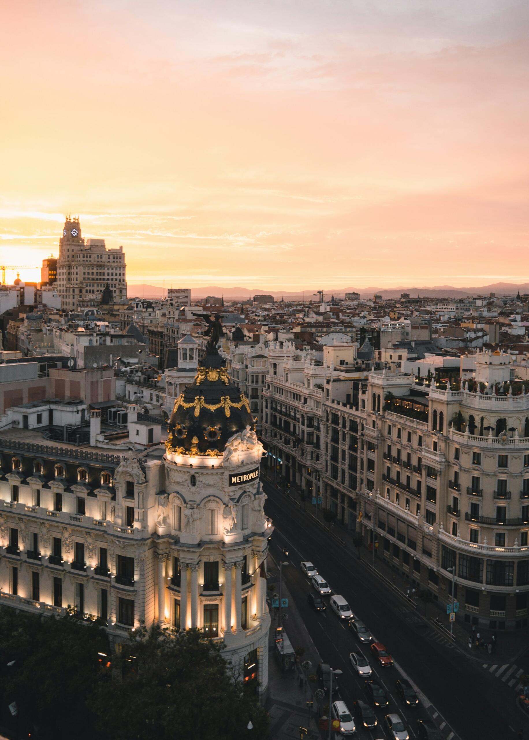 Vista aérea de Madrid al atardecer, en la que destaca la ornamentada cúpula del edificio Metropolis, iluminada. Los tonos cálidos y las calles bulliciosas crean una escena urbana llena de vida.