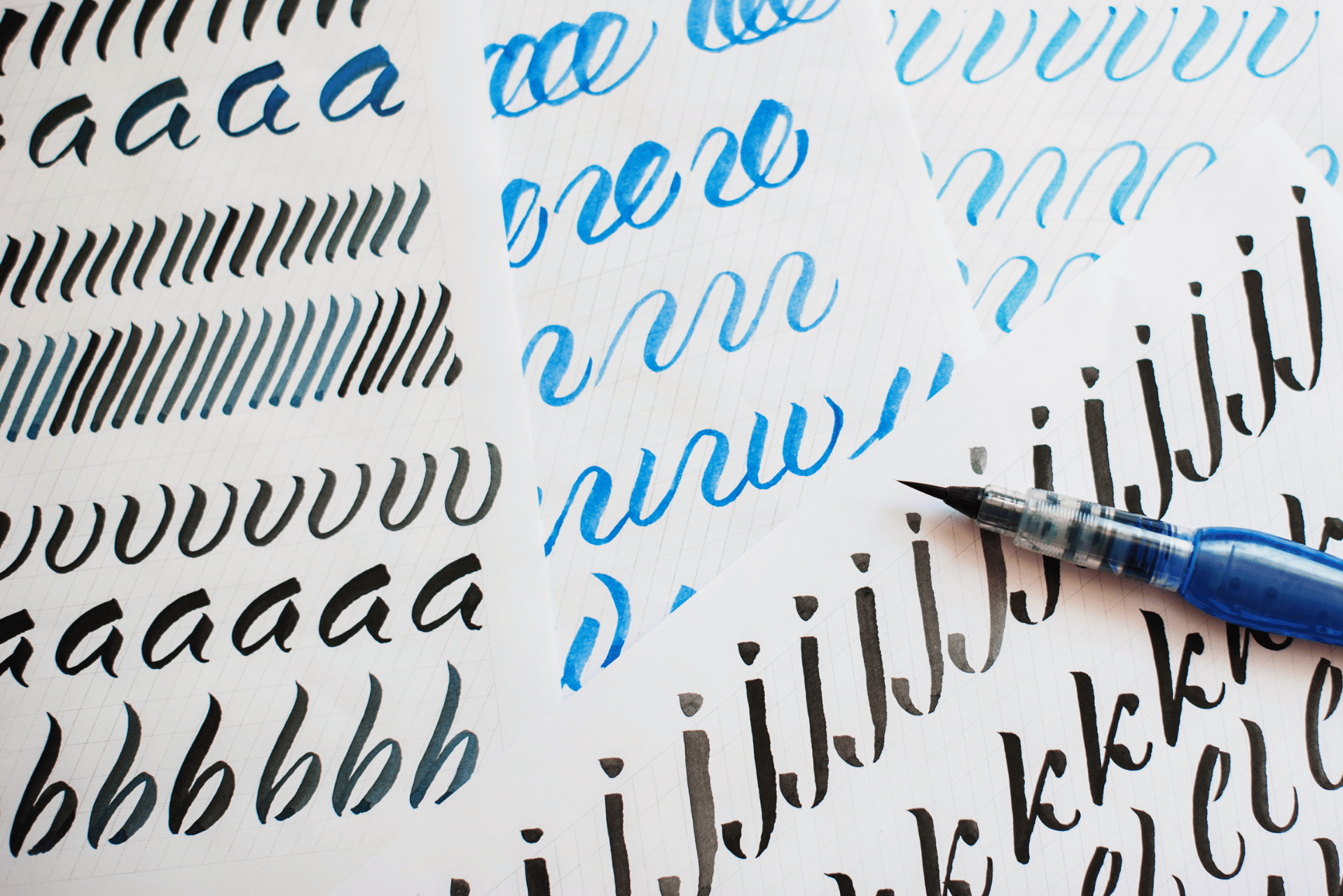 Hojas de práctica de caligrafía con diversas letras negras y azules junto a un bolígrafo.