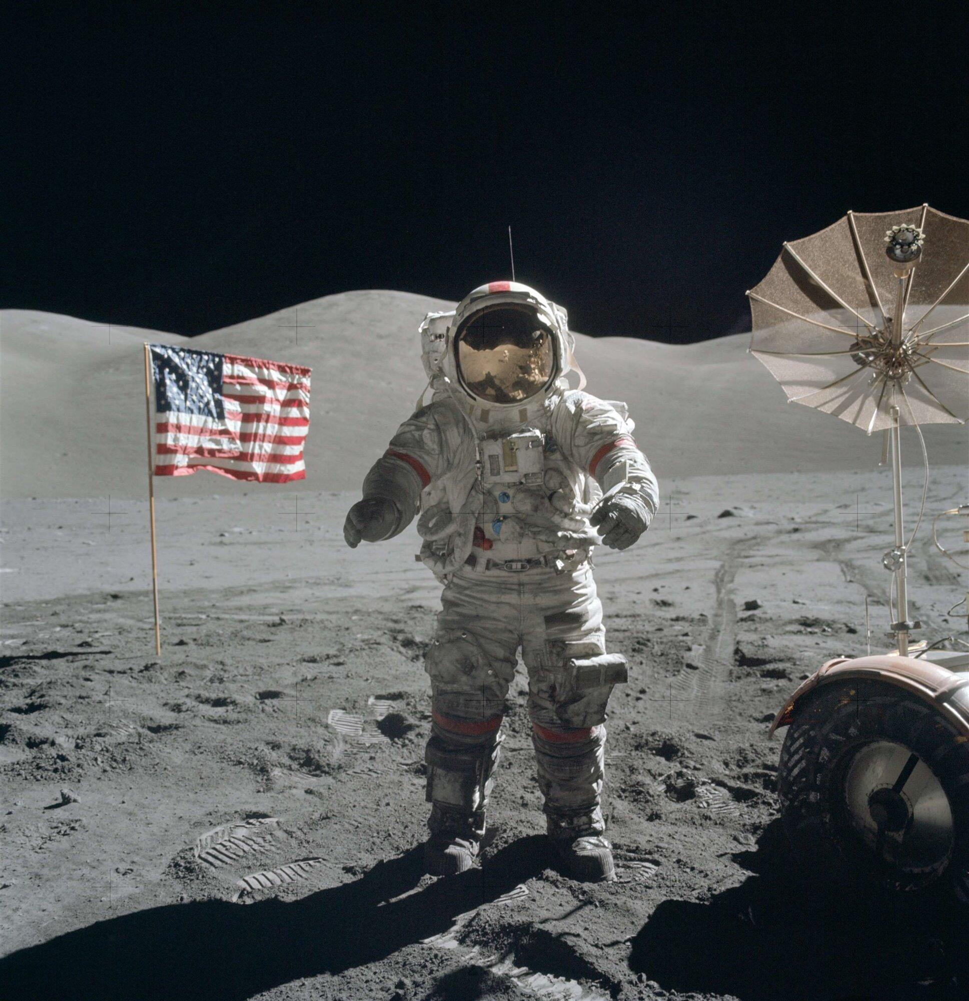 Un astronauta en la superficie lunar posa junto a una bandera estadounidense. Se divisan un vehículo lunar y una antena, con un paisaje árido y gris bajo un cielo negro.