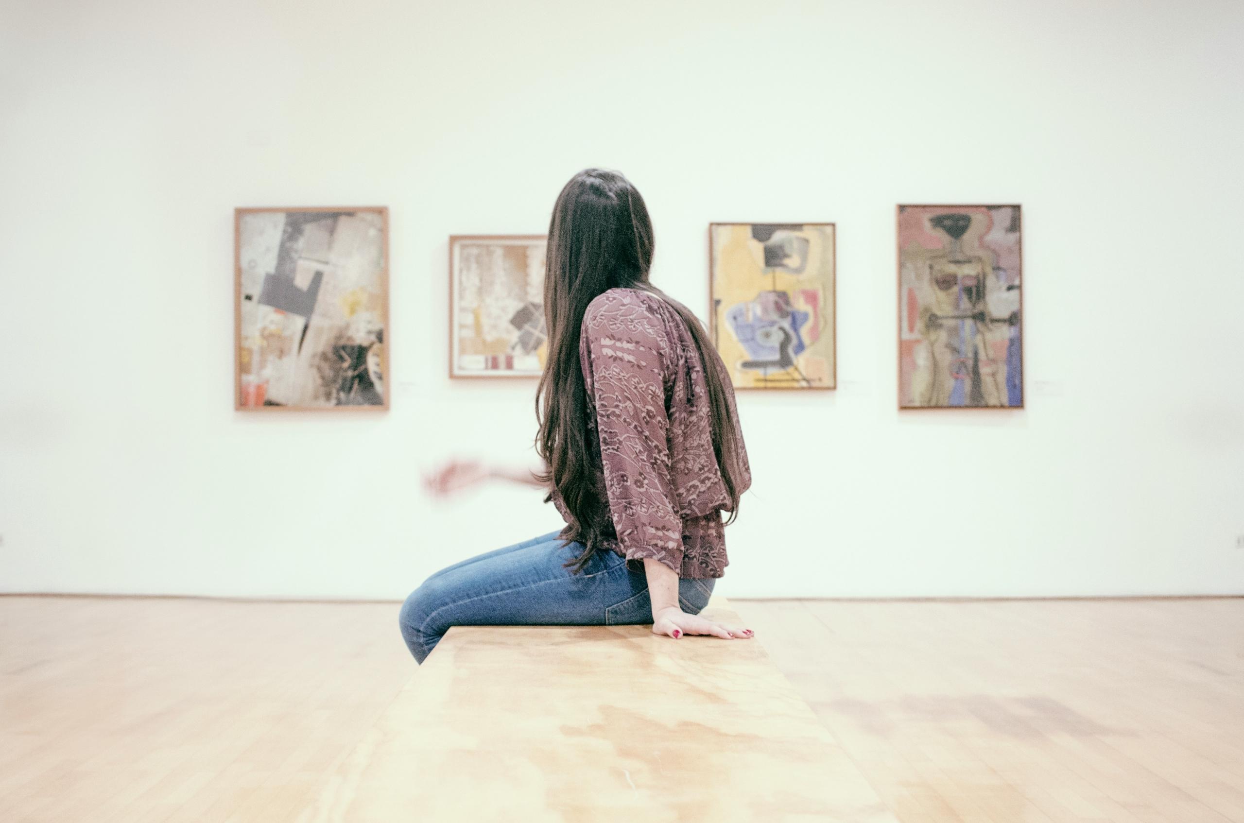 Mujer sentada en un museo o galería de arte mirando cuadros abstractos en una pared blanca.