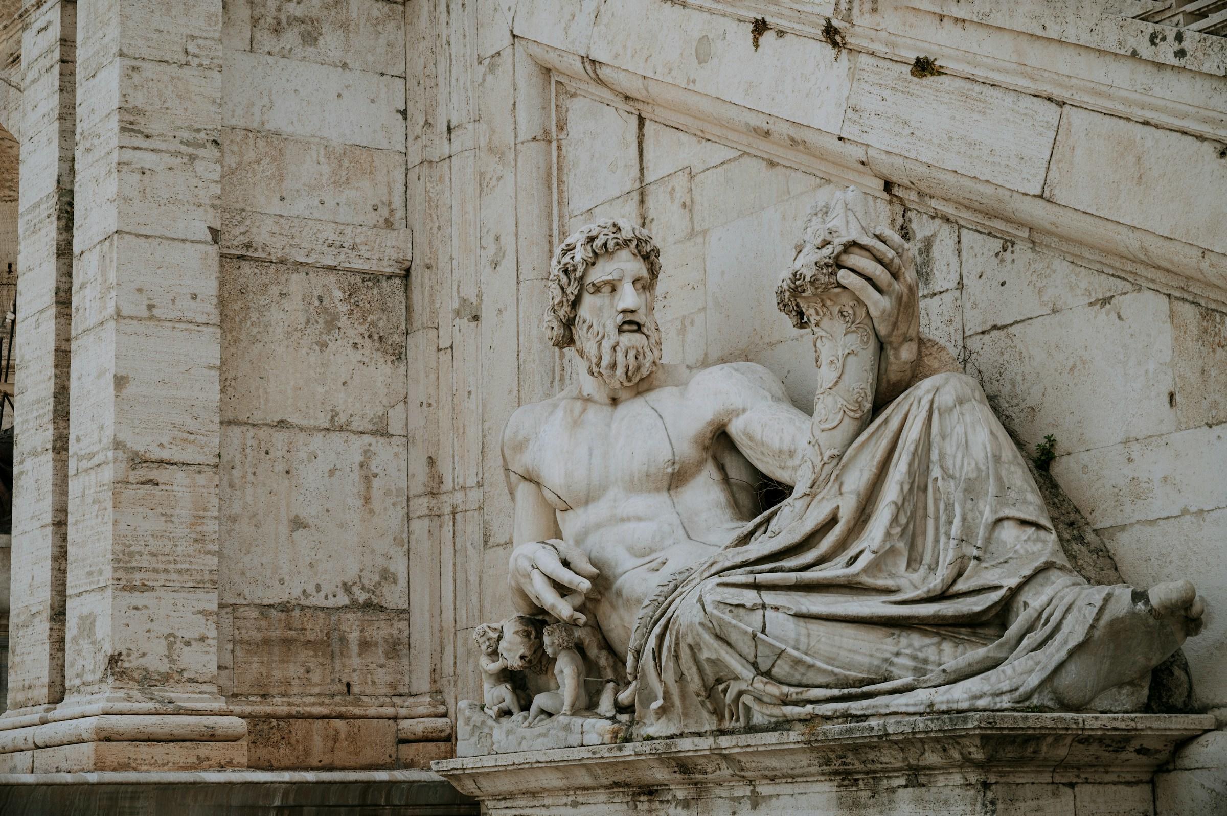 Estatua de mármol de una figura reclinada con una corona de laurel, adosada a un edificio de piedra.